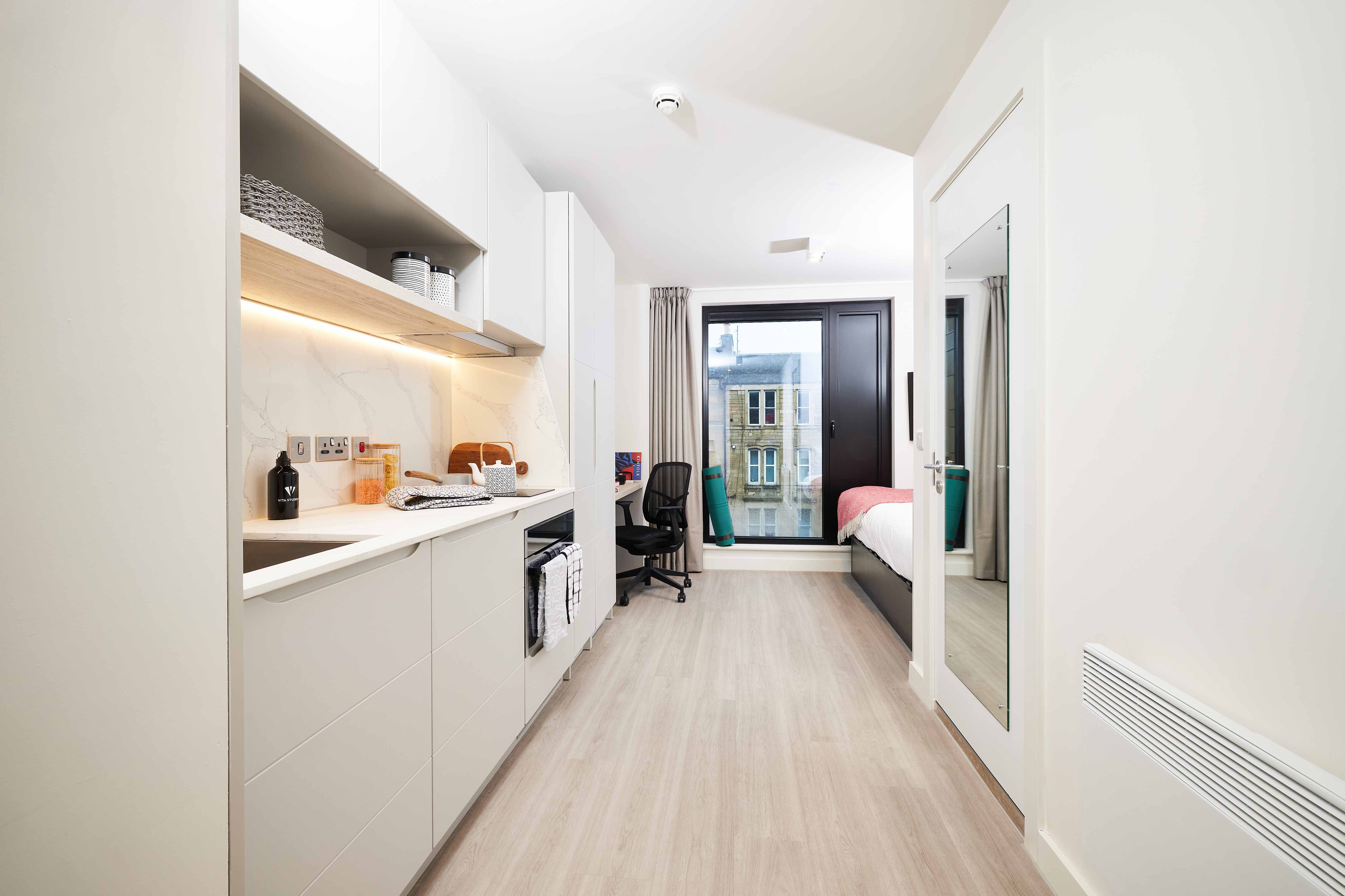 Modern studio apartment with a compact kitchen on the left, featuring white cabinetry and shelves with woven baskets. A seating area with a desk and chair is near a large window. A bed with a red bedspread is positioned on the right against the wall.