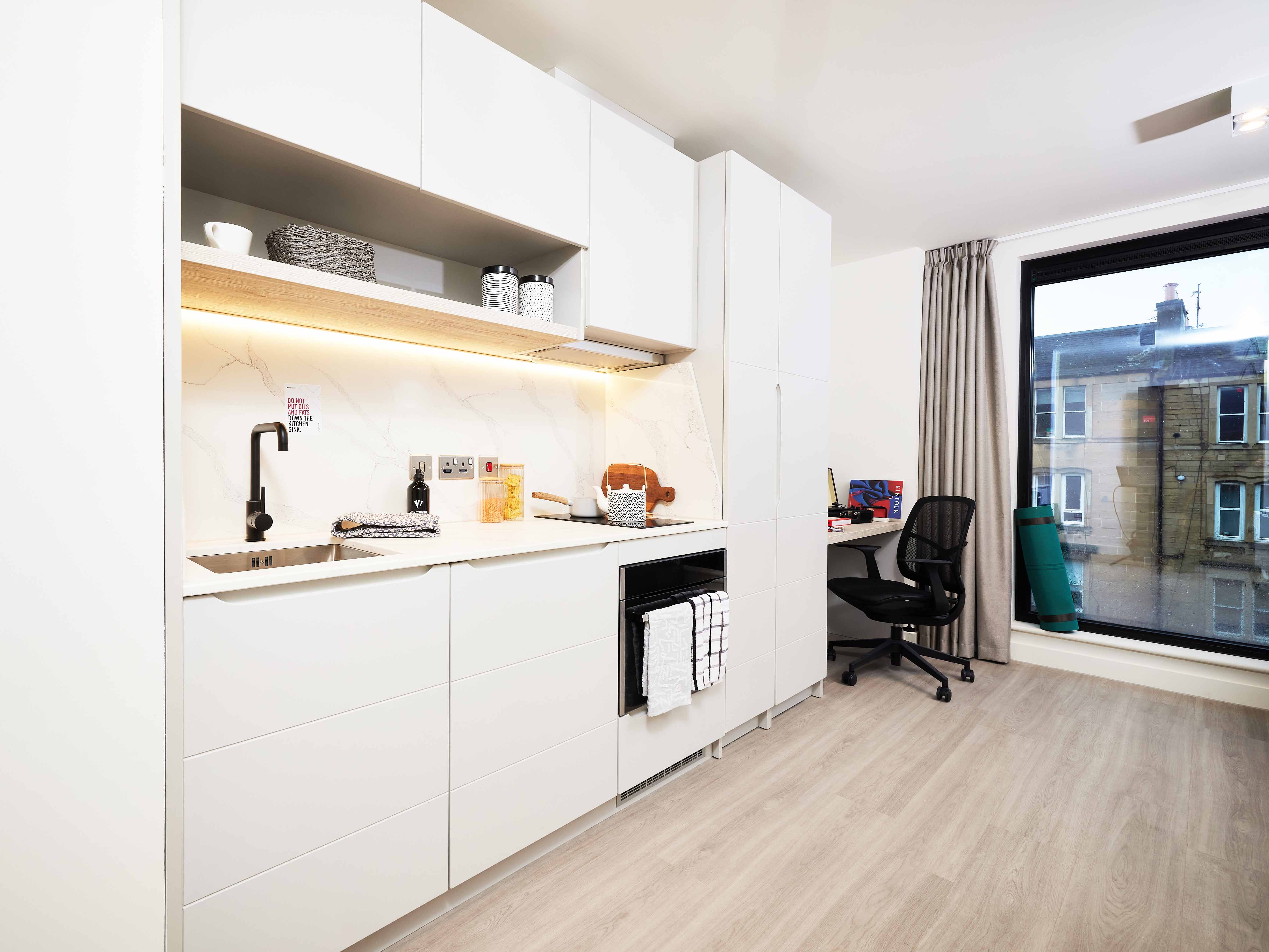 Modern kitchen in a bright room with light wood floors. White cabinets, marble backsplash, built-in oven, and sink are visible. A desk with a chair is near a large window with the view of buildings outside.