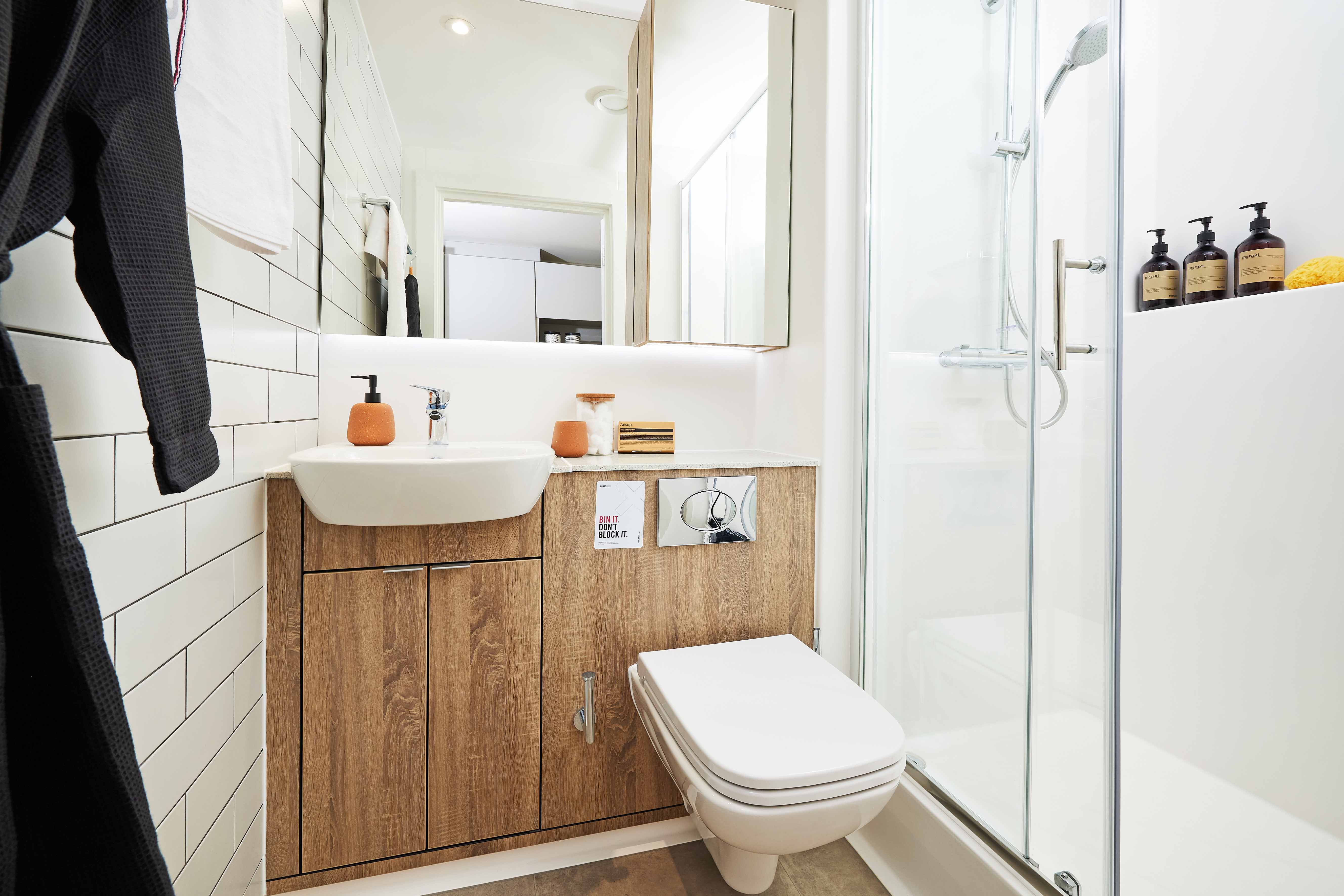 A modern bathroom with a wooden vanity holding a white sink, a wall-mounted toilet, and a glass-enclosed shower. The wall features white tiles, and there are three soap dispensers on a recessed shelf in the shower area.