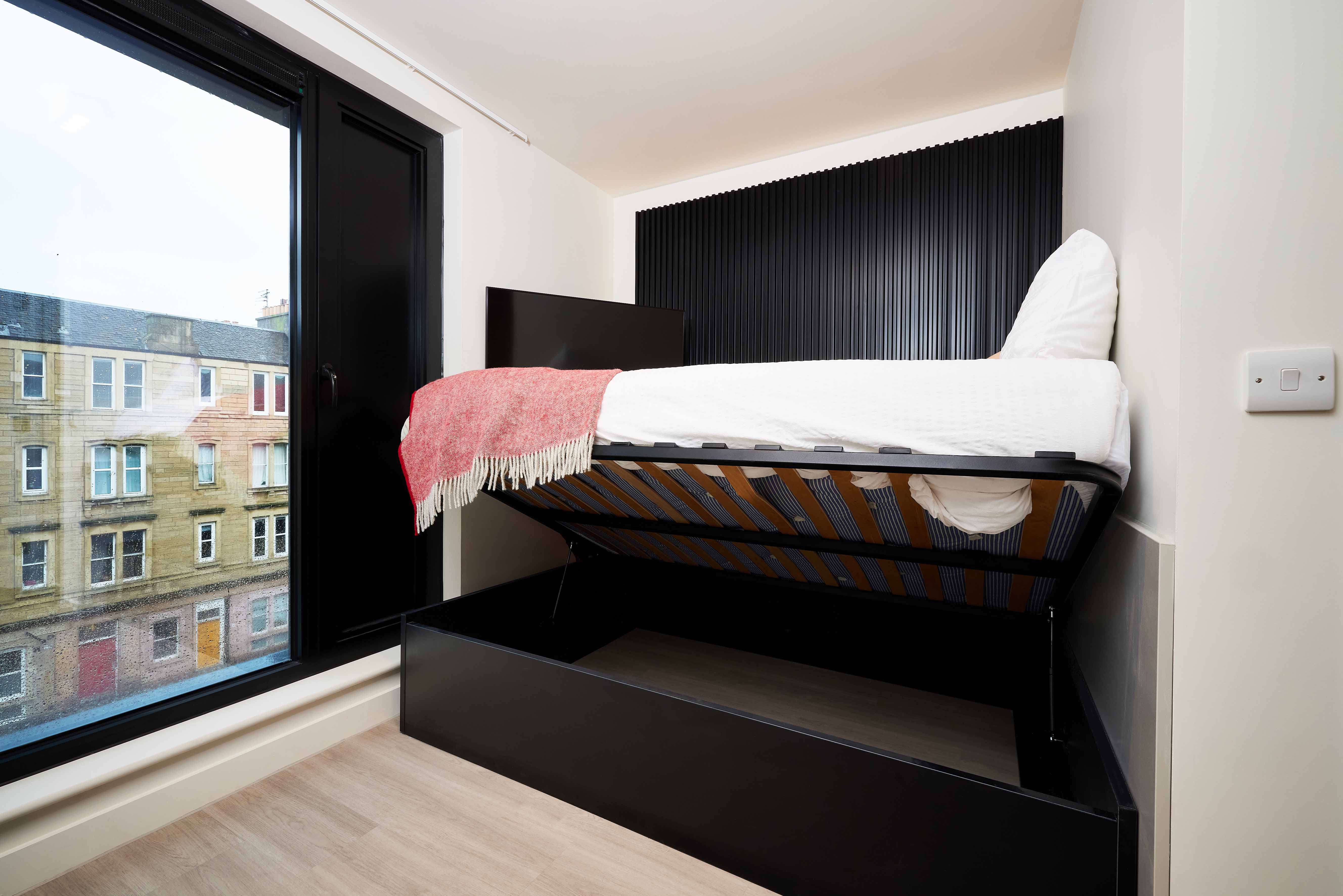 A modern studio apartment features a neatly made bed with a red throw, elevated to reveal storage underneath. Large window offers a view of neighboring buildings. Black paneling adds a sleek touch to the interior.