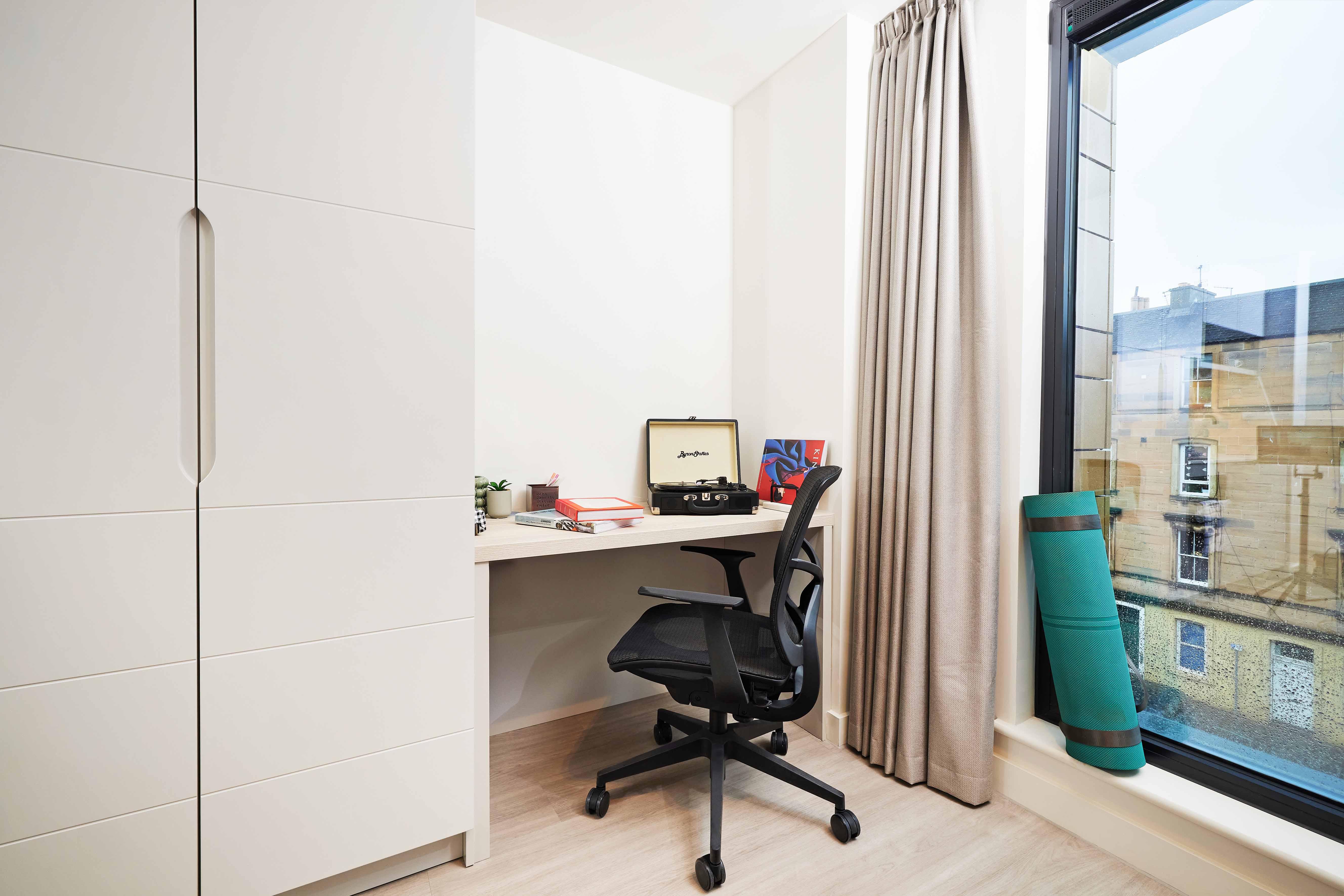 A minimalist home office with a black swivel chair at a desk. The desk has books, a small plant, and a wooden radio. Beige curtains hang by a large window with a view of buildings. A rolled yoga mat rests beside the window.