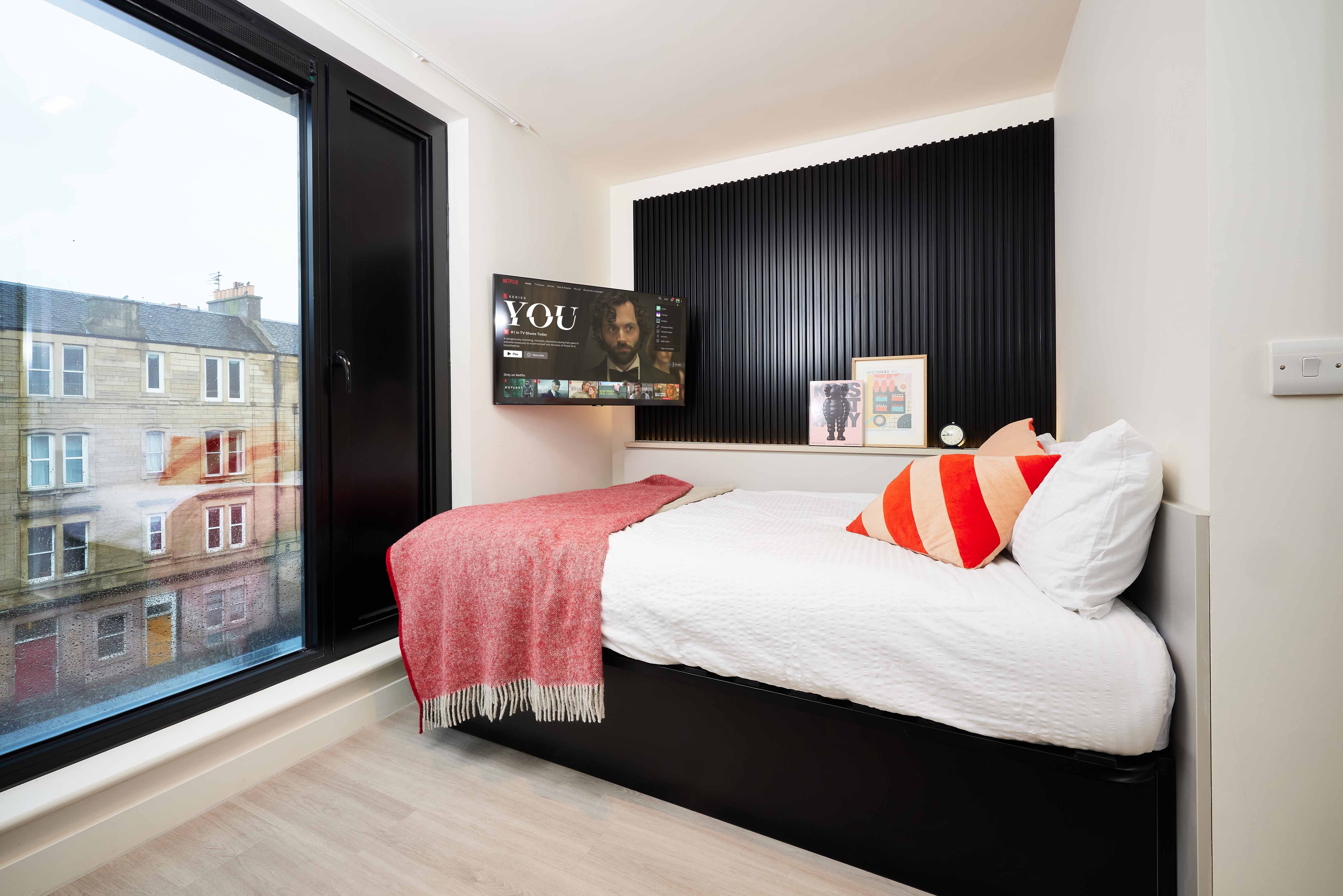 A cozy bedroom with a single bed covered in white bedspread and a red striped pillow. A red throw blanket is draped over it. A flat-screen TV is mounted on a black paneled wall. A large window shows buildings outside. A clock and books are on a shelf.