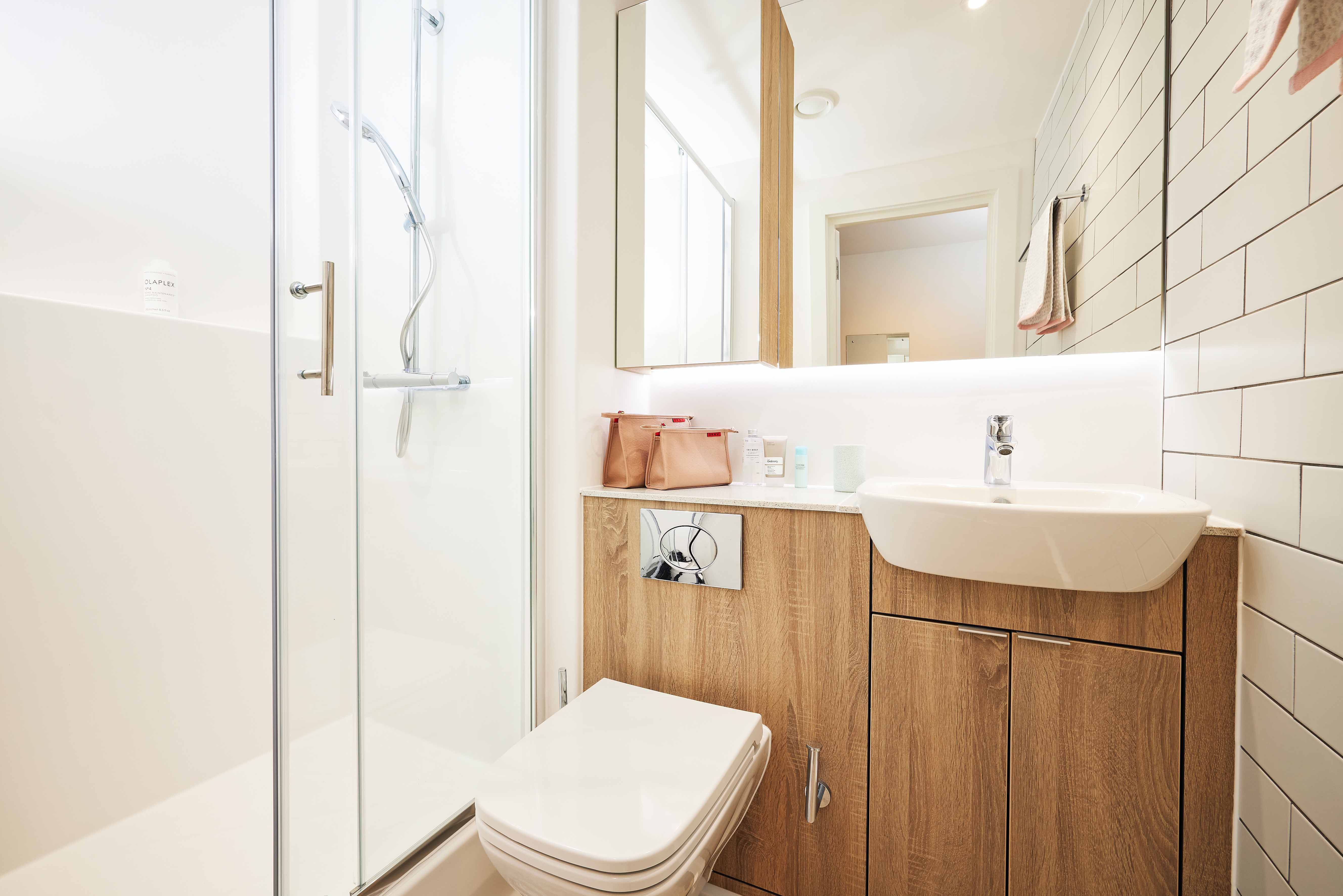 A modern bathroom with a glass-enclosed shower, white sink, and wooden cabinet. A toilet is adjacent to the cabinet. The walls have white tiles, and toiletries are neatly arranged on the countertop. Bright, natural lighting fills the space.