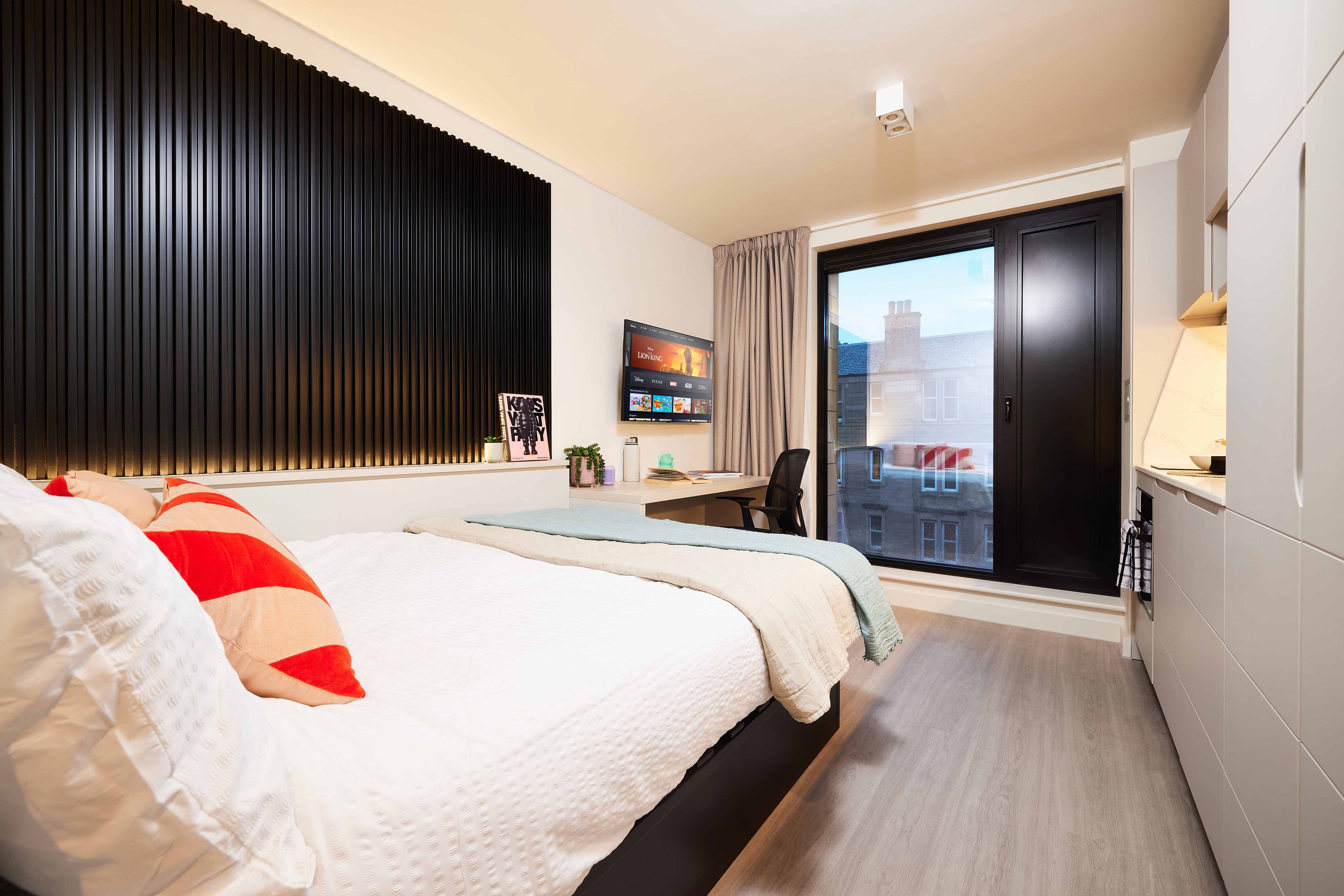 Modern bedroom with a neatly made bed featuring a red and beige pillow. A desk with a computer is placed by a window with a view of buildings. The room has a black accent wall and beige curtains, creating a cozy and stylish atmosphere.