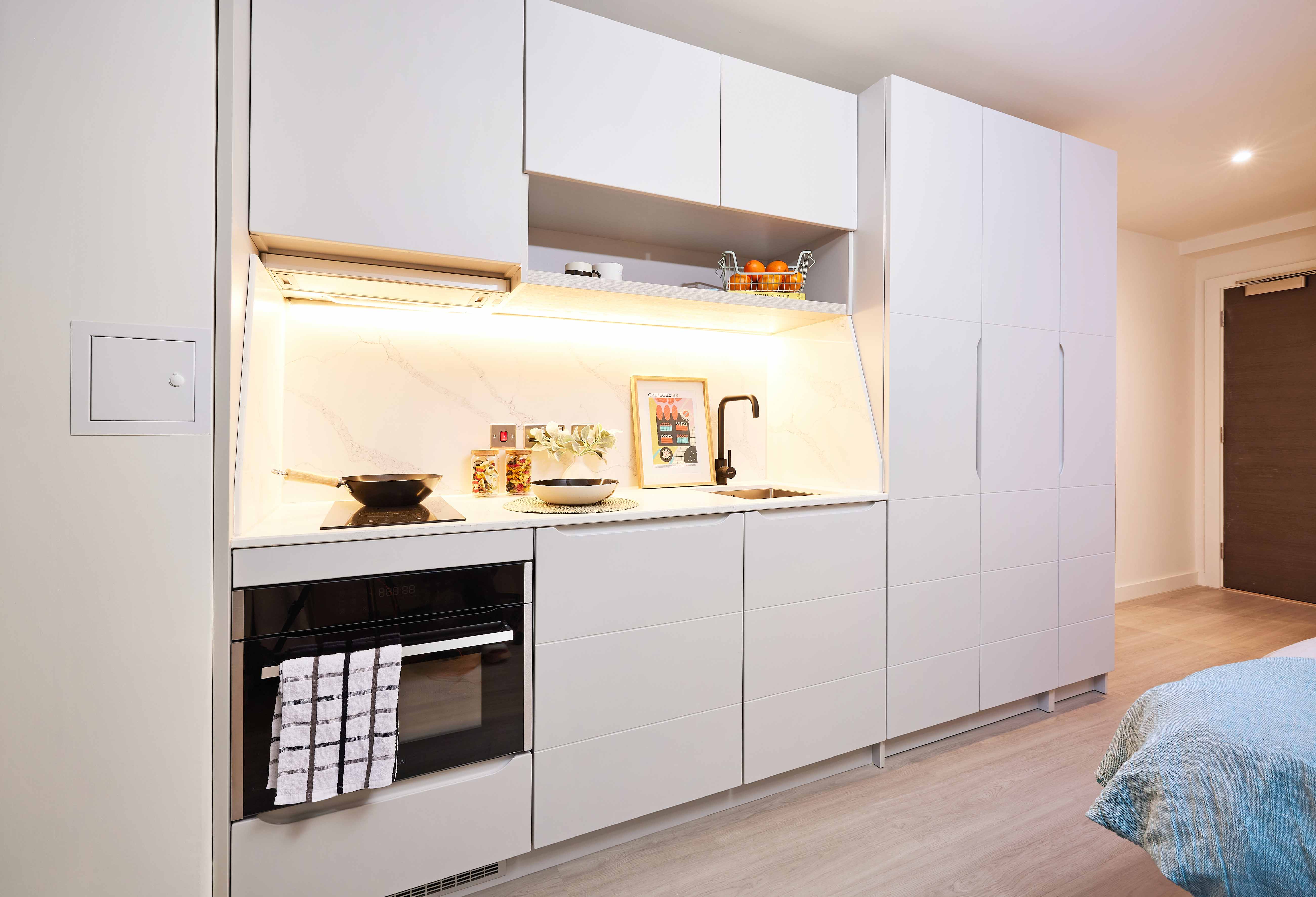 Modern kitchen with white cabinets, an electric stovetop with a pot, and an oven below. A shelf above holds a bowl of oranges. Bright lighting under the cabinets highlights a marble backsplash. There is a wooden door to the right.