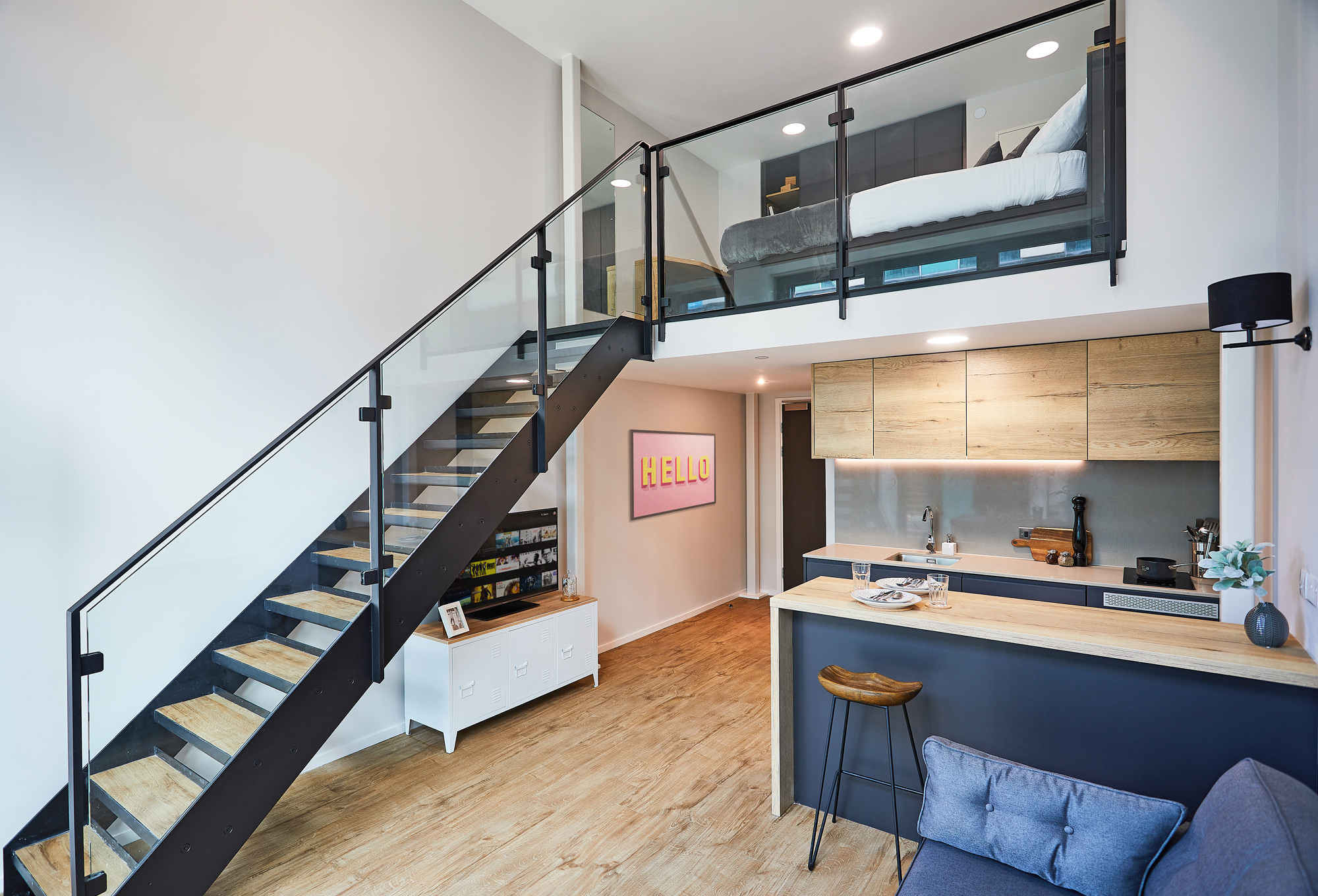 Modern loft apartment on Portland Crescent with a wooden staircase, glass railing, upper-level bedroom, and open kitchen below. Light wood floors, minimalist decor, "HELLO" wall art, and a small counter with barstools are visible.