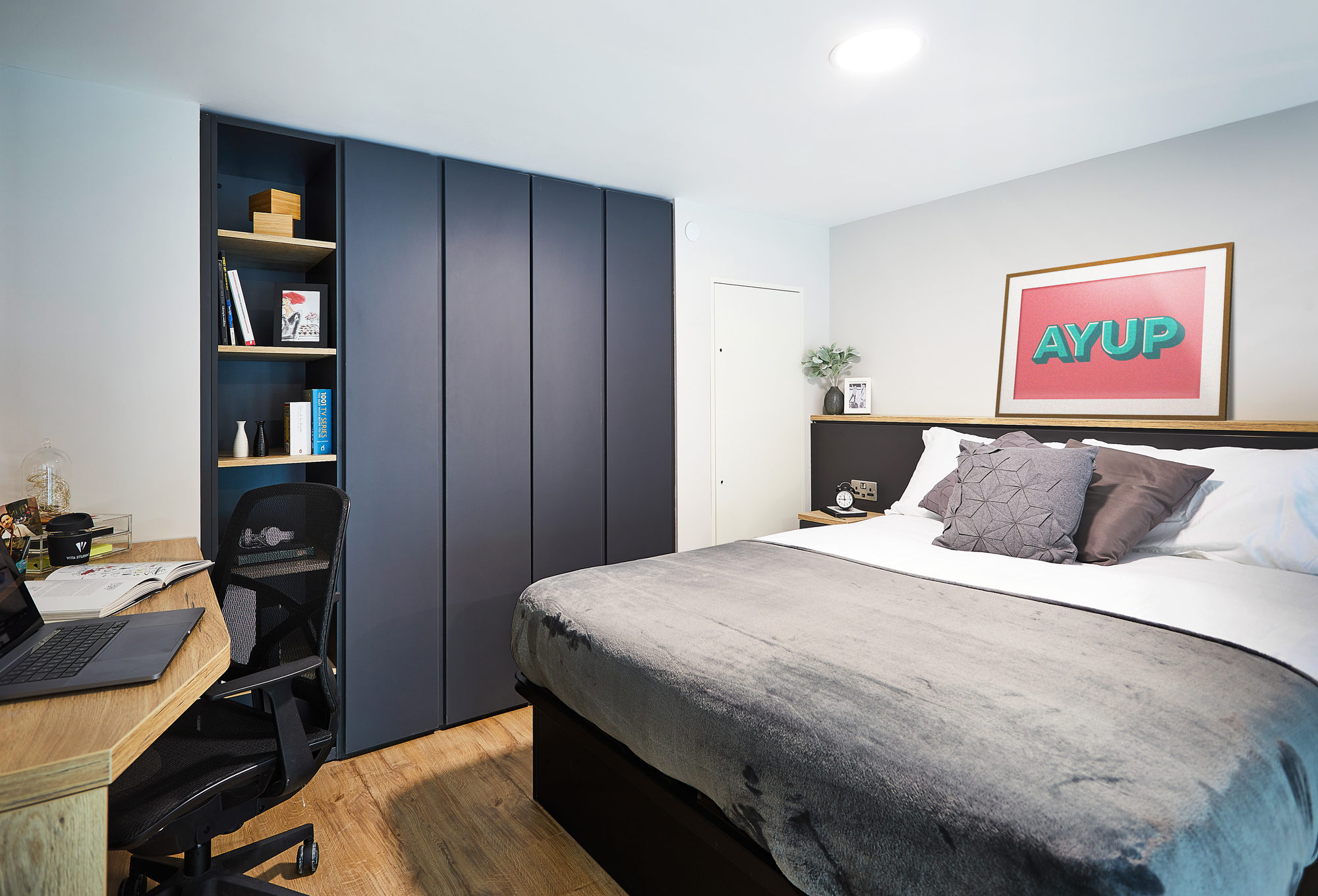 Modern bedroom at Portland Crescent with a double bed, gray bedding, dark wardrobe, open shelving with books, a wooden desk with a laptop and office chair, and a framed "AYUP" print on the wall.