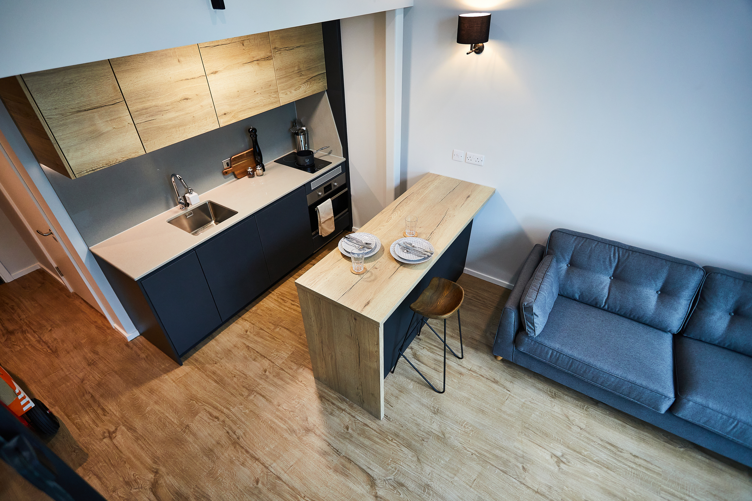 Modern apartment kitchen in Portland with wood cabinets, a sink, and built-in appliances. A wooden island with two plates and glasses is paired with a stool. A gray sofa sits to the right on wood flooring under soft Crescent lighting.