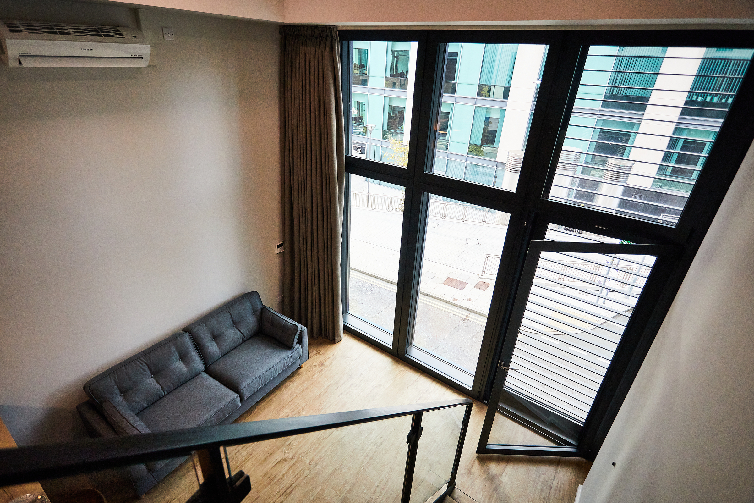 A modern living room at Portland Crescent features a gray sofa, wooden floor, large floor-to-ceiling windows, brown curtains, and a wall-mounted air conditioner, all viewed from an upper level next to a black stair railing.