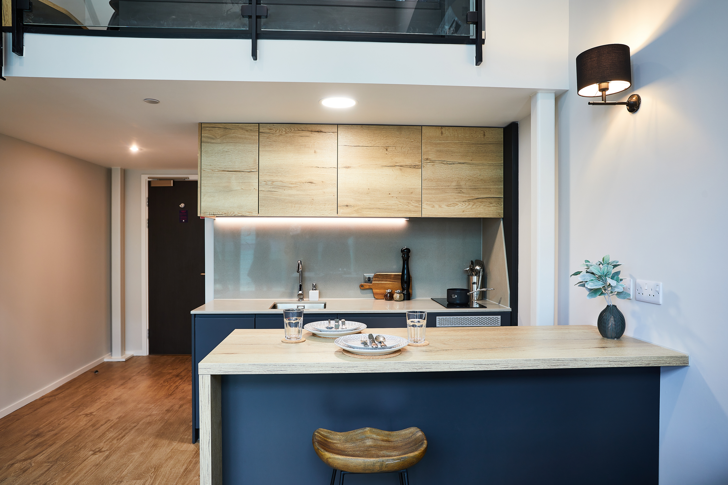 Modern kitchen at Portland Crescent with wood cabinets, a light wood countertop, blue base cabinets, and bar stools. Two plates, glasses, and a plant sit on the counter, complemented by minimalistic decor and bright lighting.