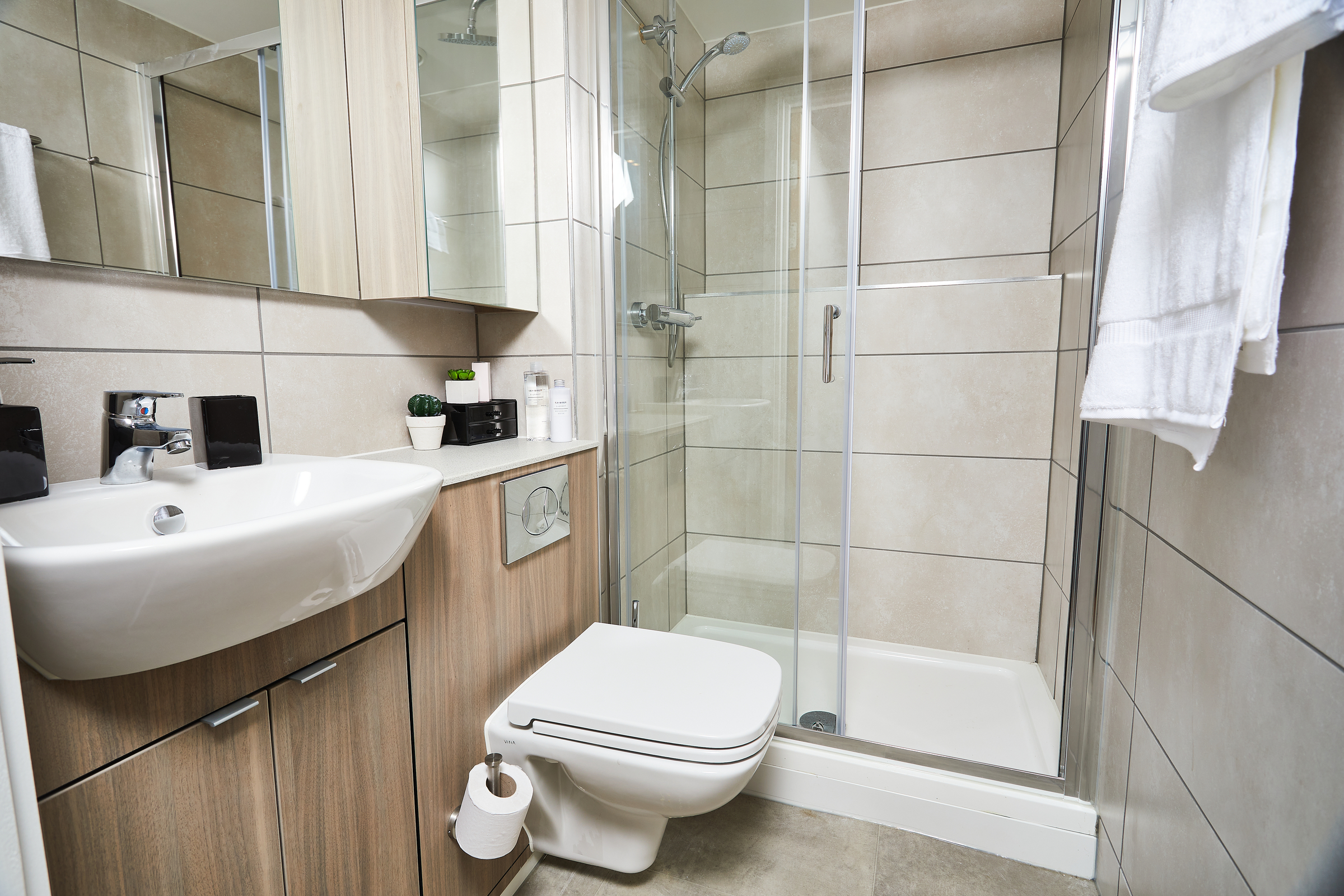 Modern bathroom at Portland Crescent with tan tiles, wall-mounted sink, white toilet, glass-enclosed shower, large mirror, wooden cabinetry, toilet paper holder, and white towels hanging on the wall.