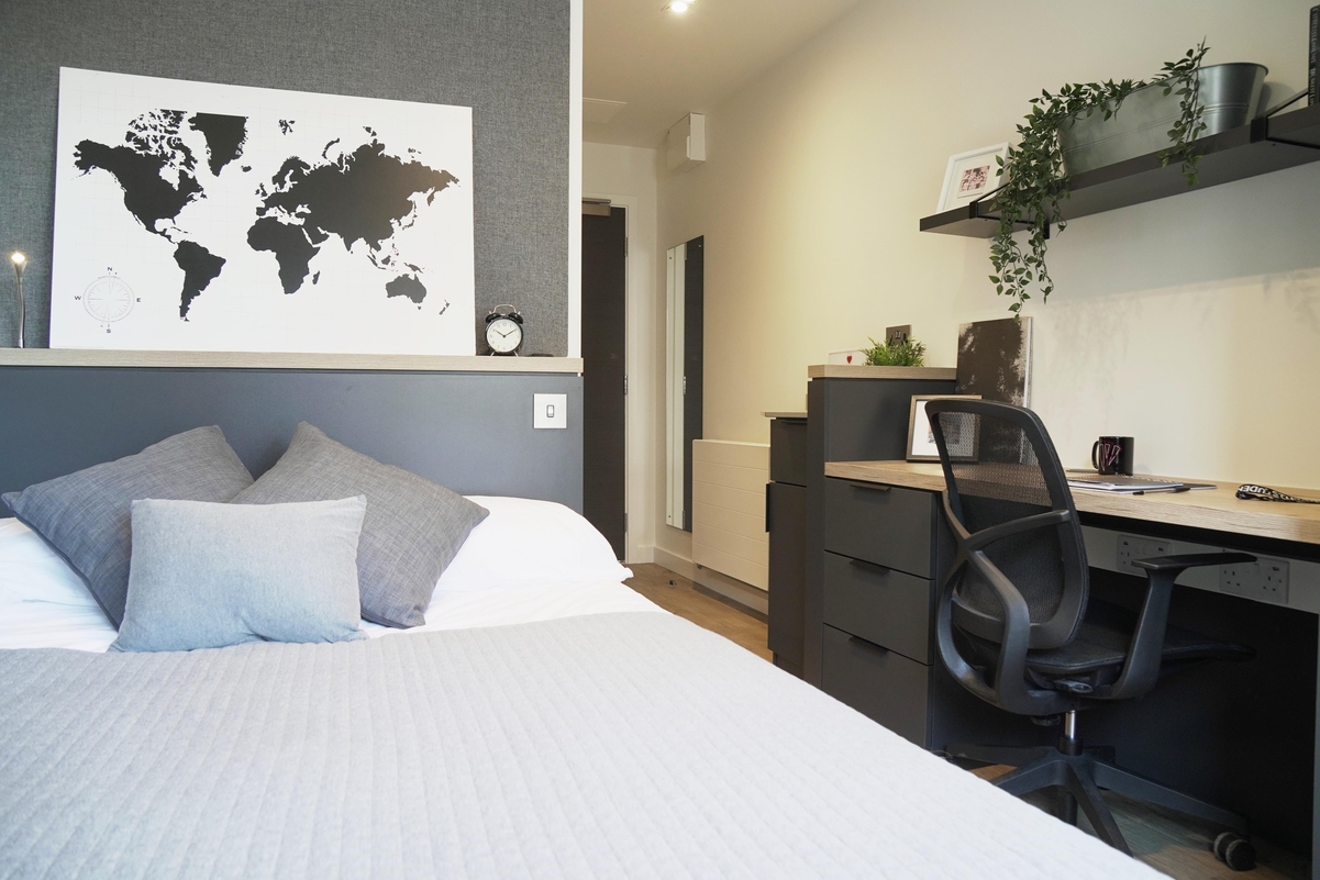 A modern, tidy Lewisham apartment bedroom with a bed in the foreground, three gray pillows, a world map on the wall, and a desk with a black chair, drawers, plants, and a mug in the background.