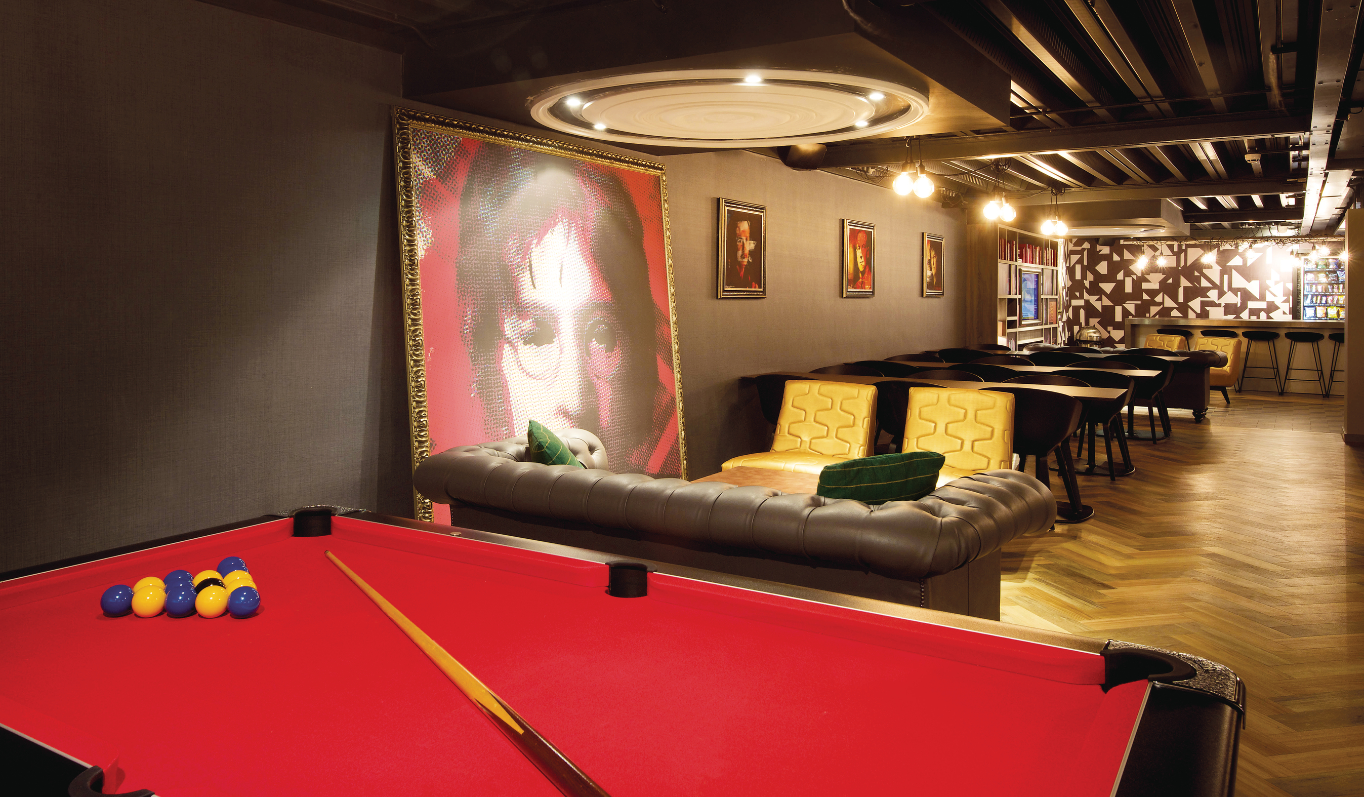 A cozy lounge in a luxury student accommodation features a red billiards table upfront. Behind it, leather couches and framed art adorn the walls. The bar area stands against a patterned wall, bathed in warm lighting that creates an inviting atmosphere for both study and relaxation.