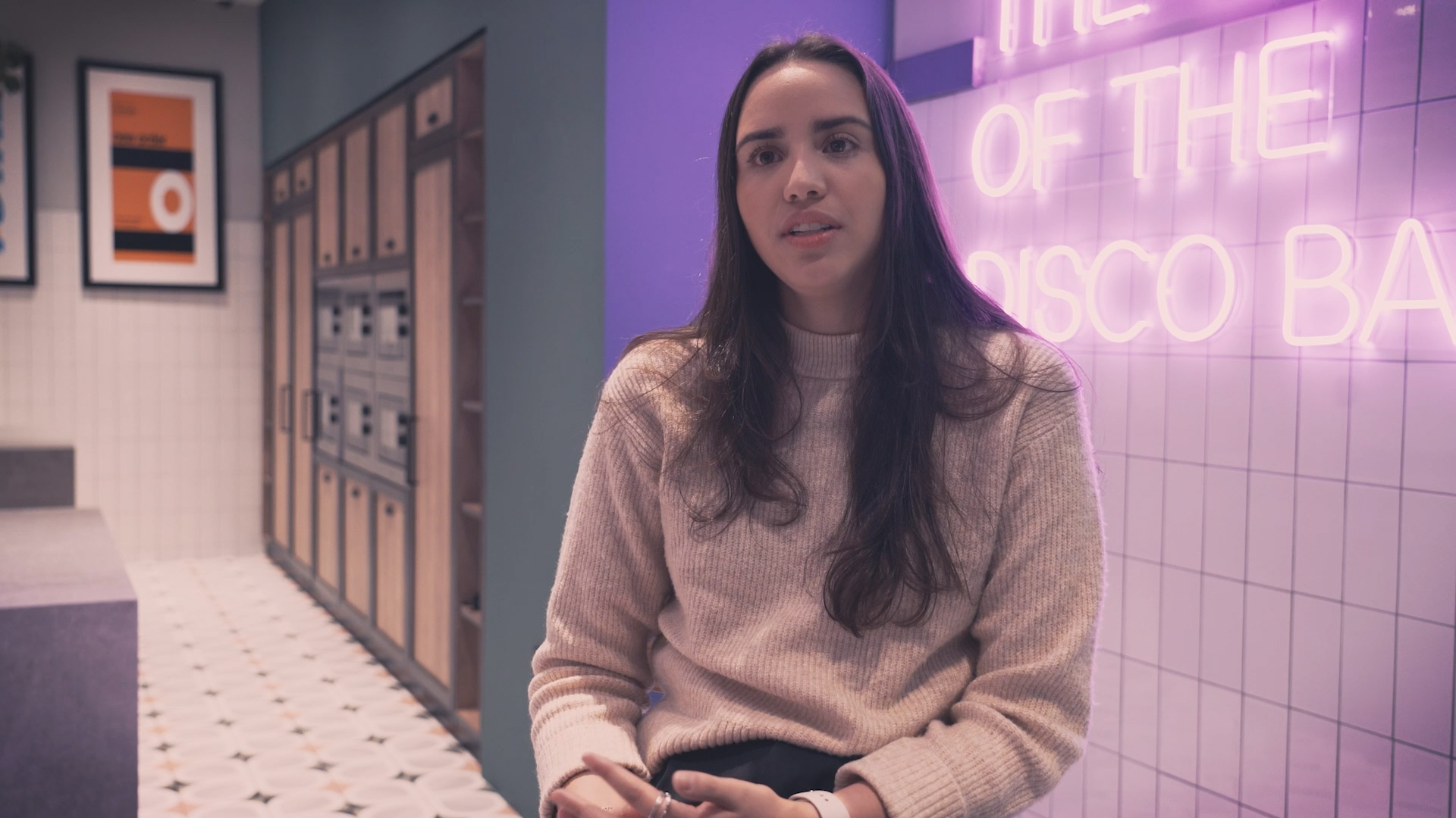 A woman with long dark hair sits in a sleek hallway of student accommodation in Coventry. She's wearing a light sweater and dark pants, with lockers to her left. Behind her, a purple wall features a neon sign that partially reads "THE OF THE DISCO BAR.