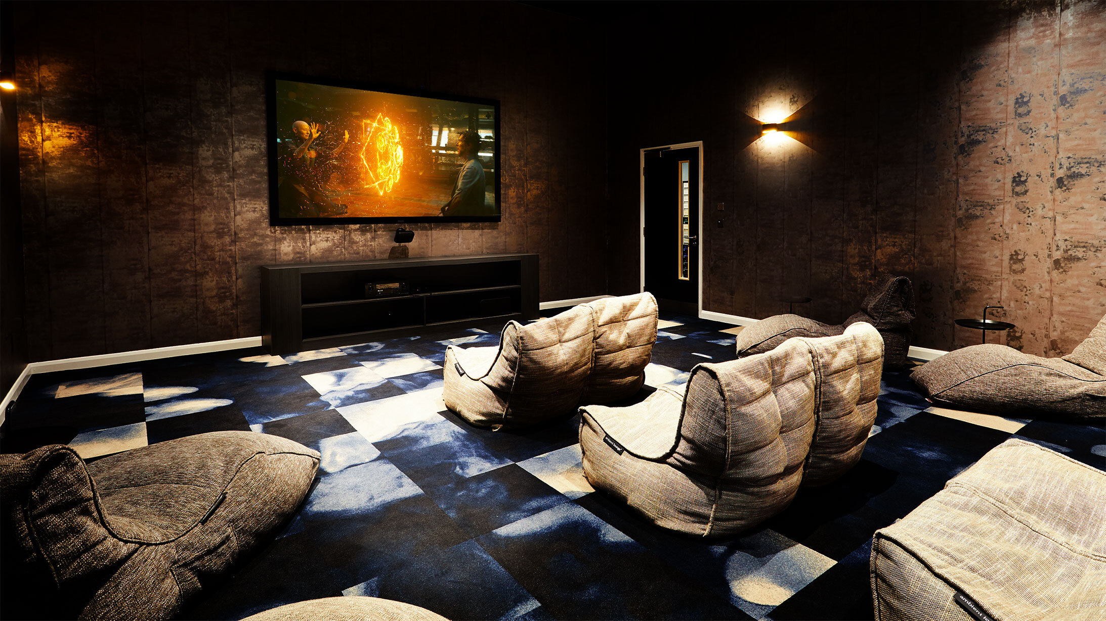 A home theater room features several plush bean bag chairs facing a wall-mounted TV displaying a movie scene, with dim lighting casting soft shadows on the dark textured walls. A patterned blue and white carpet adds charm, while square accents subtly complement the ambiance.