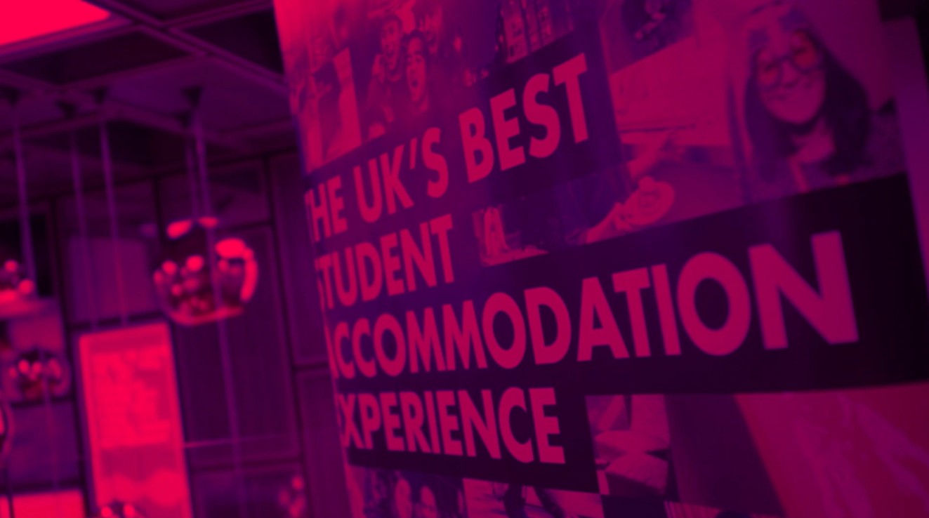 A pink-tinted banner reads "THE UK'S BEST STUDENT ACCOMMODATION EXPERIENCE," with blurred images of people in the background, and ceiling lights reflected on glass in a circle and square pattern.