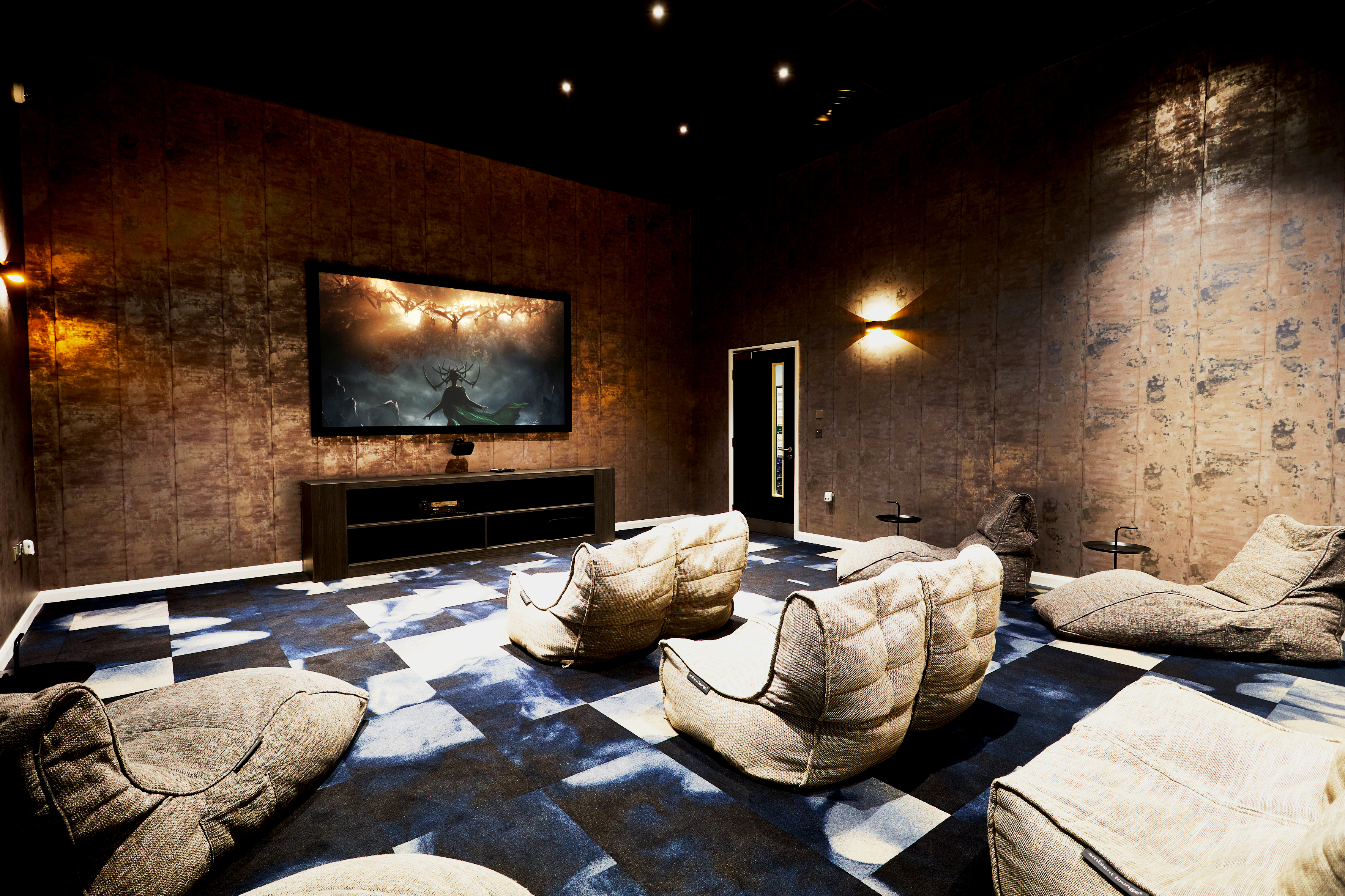 A modern home theater room with cushioned seats on a blue and white carpet features a large screen displaying an action scene. Walls are dark with subtle patterns, and soft lighting accents the space, creating a cozy atmosphere. Circular light fixtures add an elegant touch.