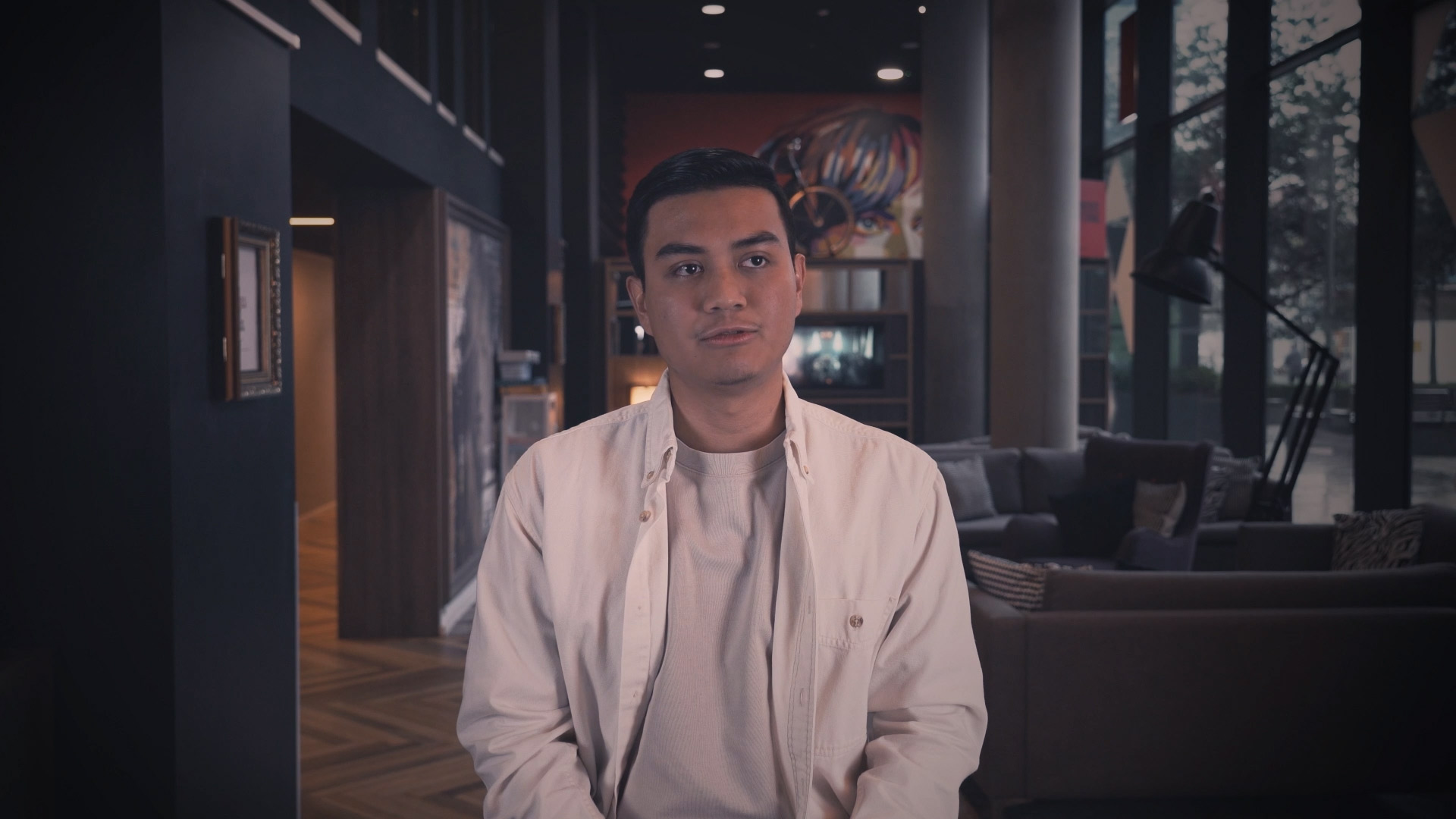 A man in a white shirt is seated indoors within a modern, warmly-lit student accommodation in Coventry, complete with stylish artwork, comfortable furniture, and expansive windows.