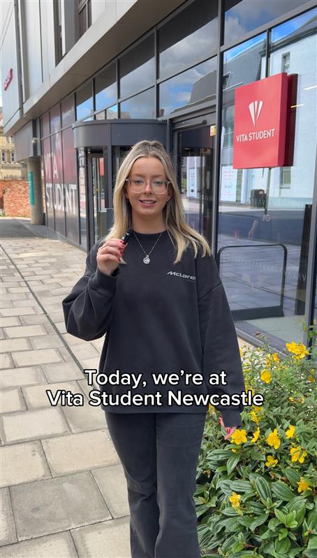 A person in a black sweatshirt and glasses stands outside Vita Student Newcastle. The building is modern with glass windows. Yellow flowers are in the foreground. Text on the image reads, "Today, we're at Vita Student Newcastle.