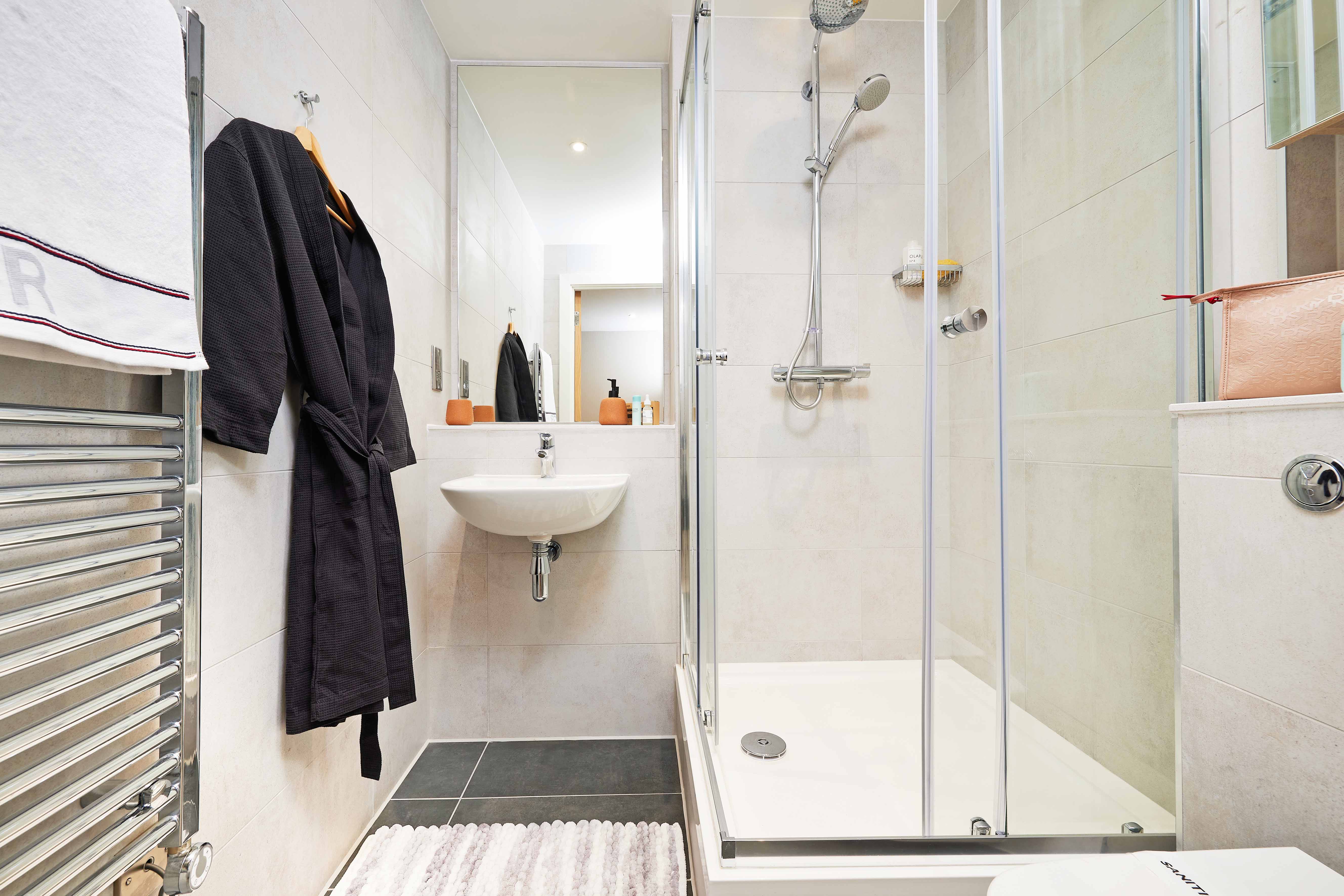 A modern bathroom with a glass shower enclosure, white sink, and wall-mounted faucet. A black robe hangs near a heated towel rail with white towels. The floor has a striped rug, and there are soap dispensers on the sink and shower shelf.