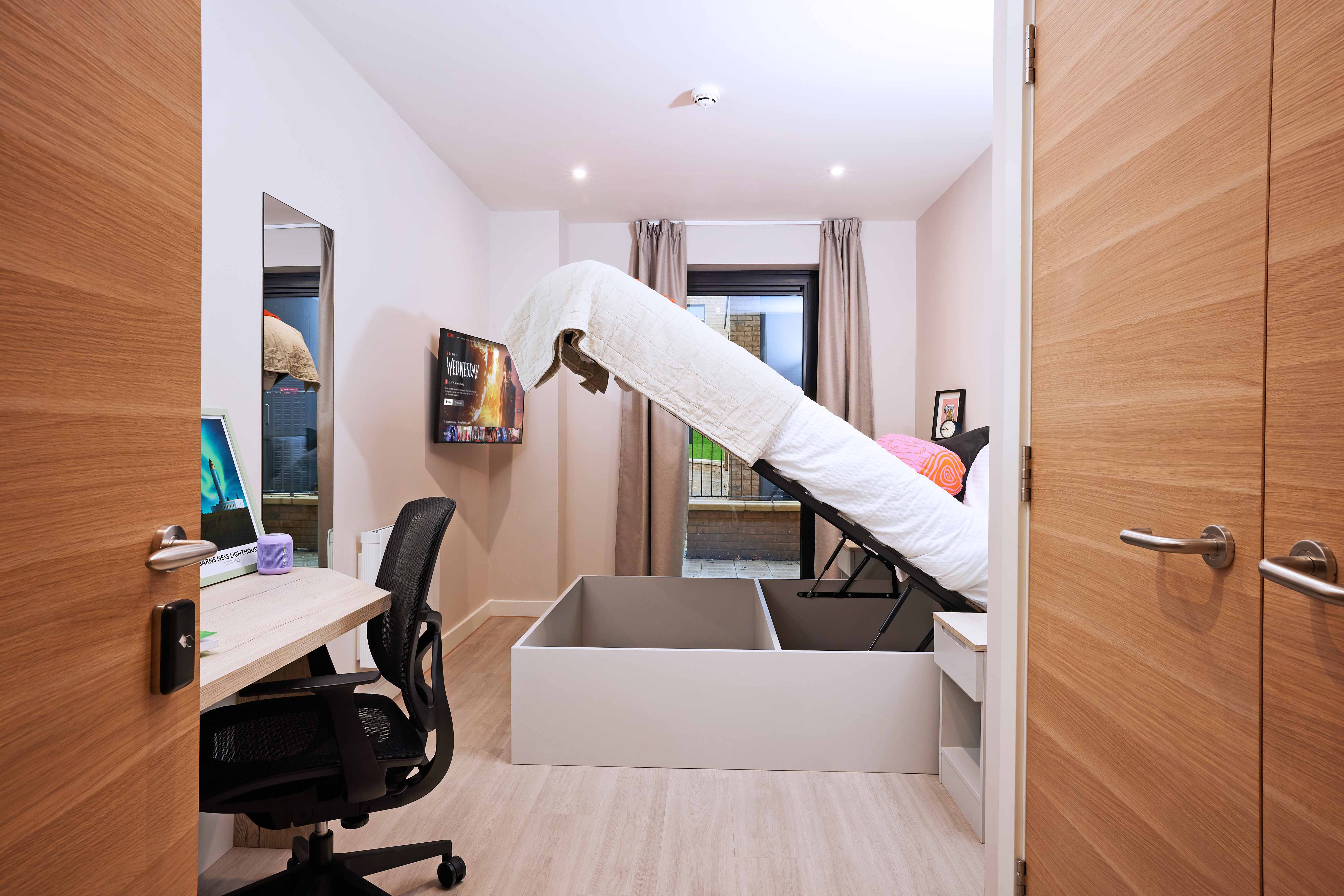 A small bedroom with a lifted bed revealing storage underneath. There's a desk with a computer and a chair on the left, a wall-mounted TV, and a window with closed curtains. Walls and doors are wood-toned, and the floor is light-colored.