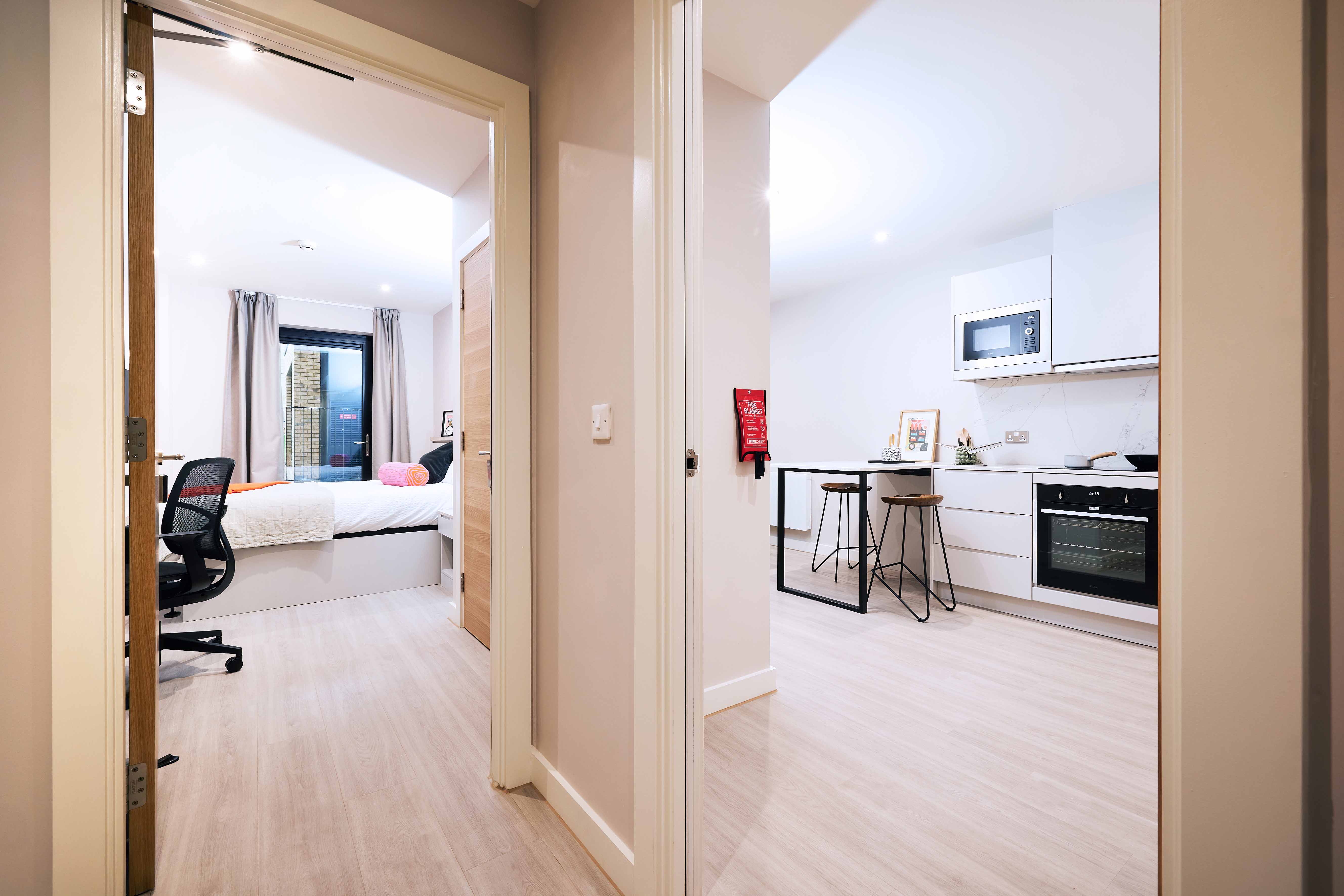 A modern apartment hallway with a view into a bedroom on the left and a kitchen straight ahead. The bedroom has a large bed and desk; the kitchen features white cabinets, an oven, a microwave, and a small dining area with stools.