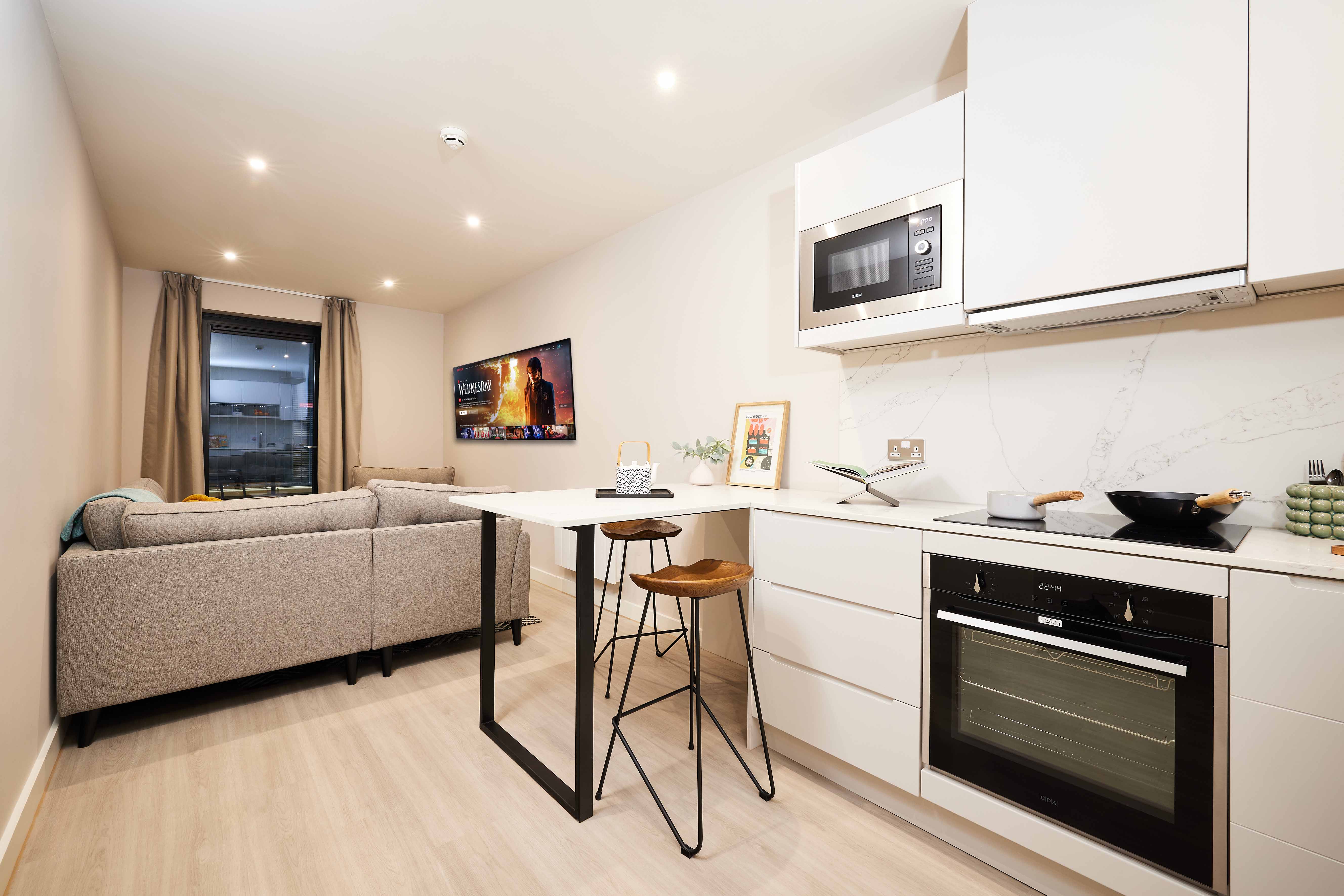Modern studio apartment with a small kitchen and living area. The kitchen has white cabinets, stove, and microwave. A table with stools divides the space. A gray couch faces a wall-mounted TV. A window with curtains is on the left wall.