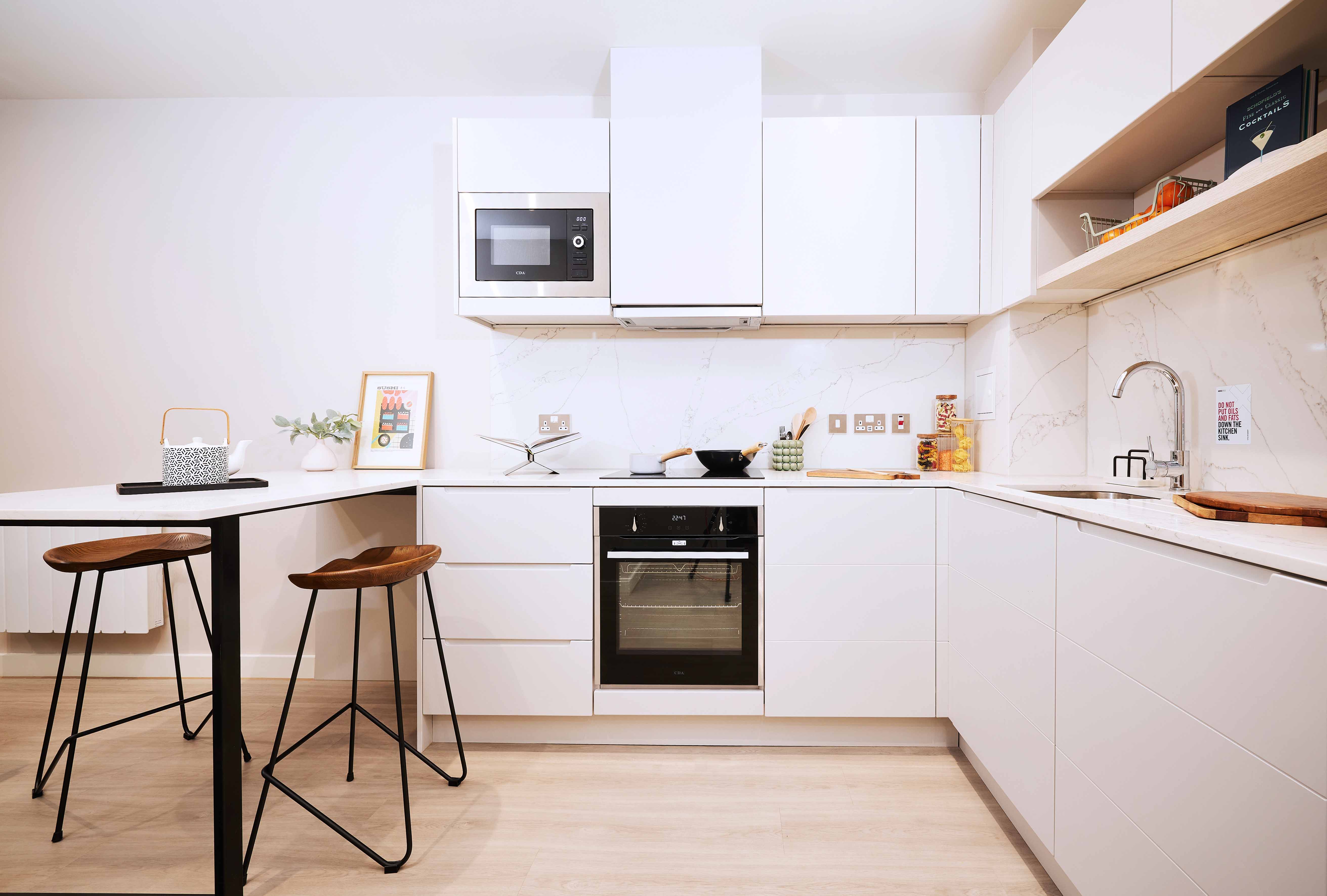 Modern kitchen with white cabinets, a built-in oven, and a microwave. A small wooden table with two stools is on the left. Countertop has a few items like plants and utensils. Light wood flooring and white walls complete the minimalist look.