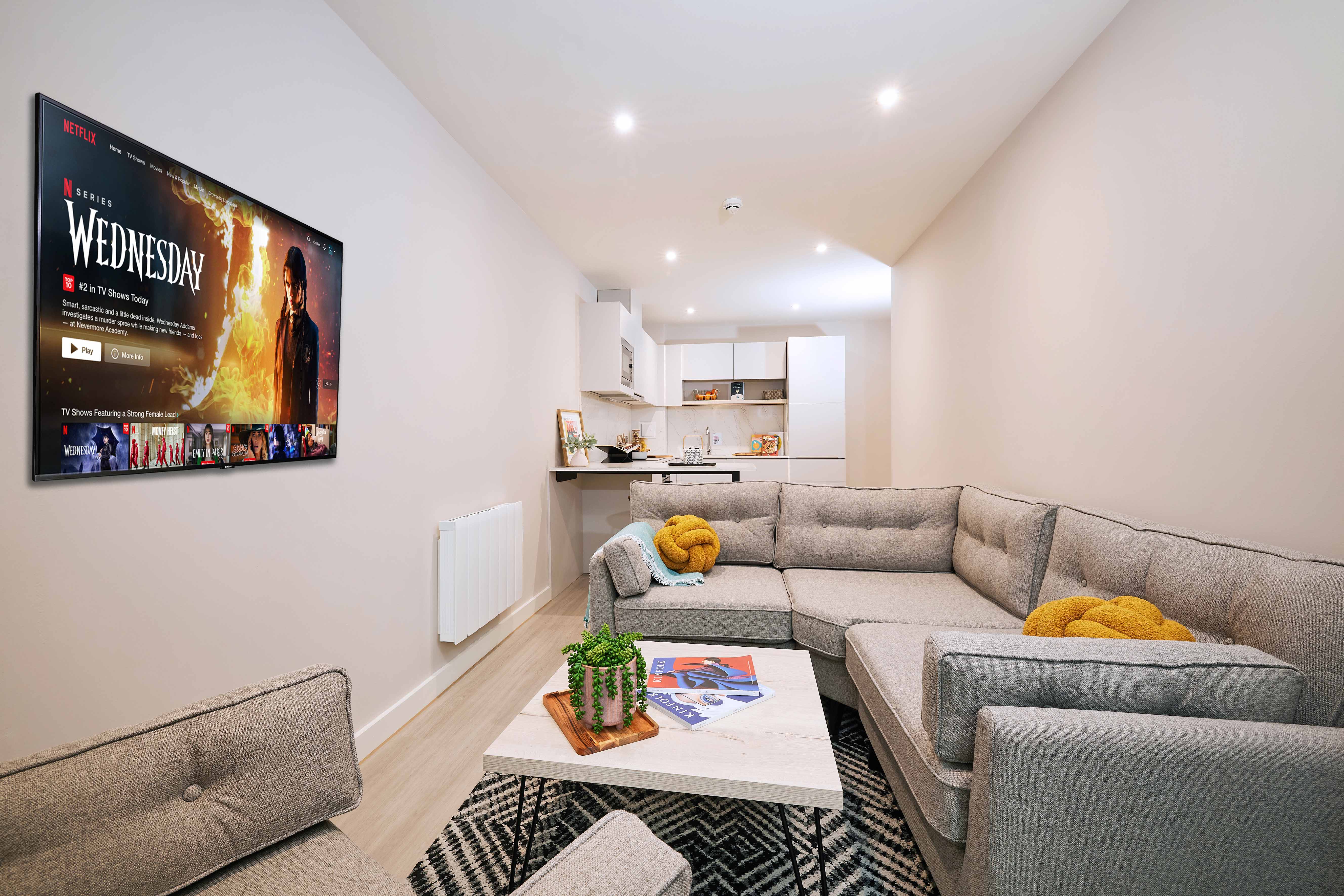 A modern living room with a gray sectional sofa, yellow cushions, and a geometric rug. A flat-screen TV on the wall displays a show. In the background, there's a small kitchen with white cabinets and a wooden counter.