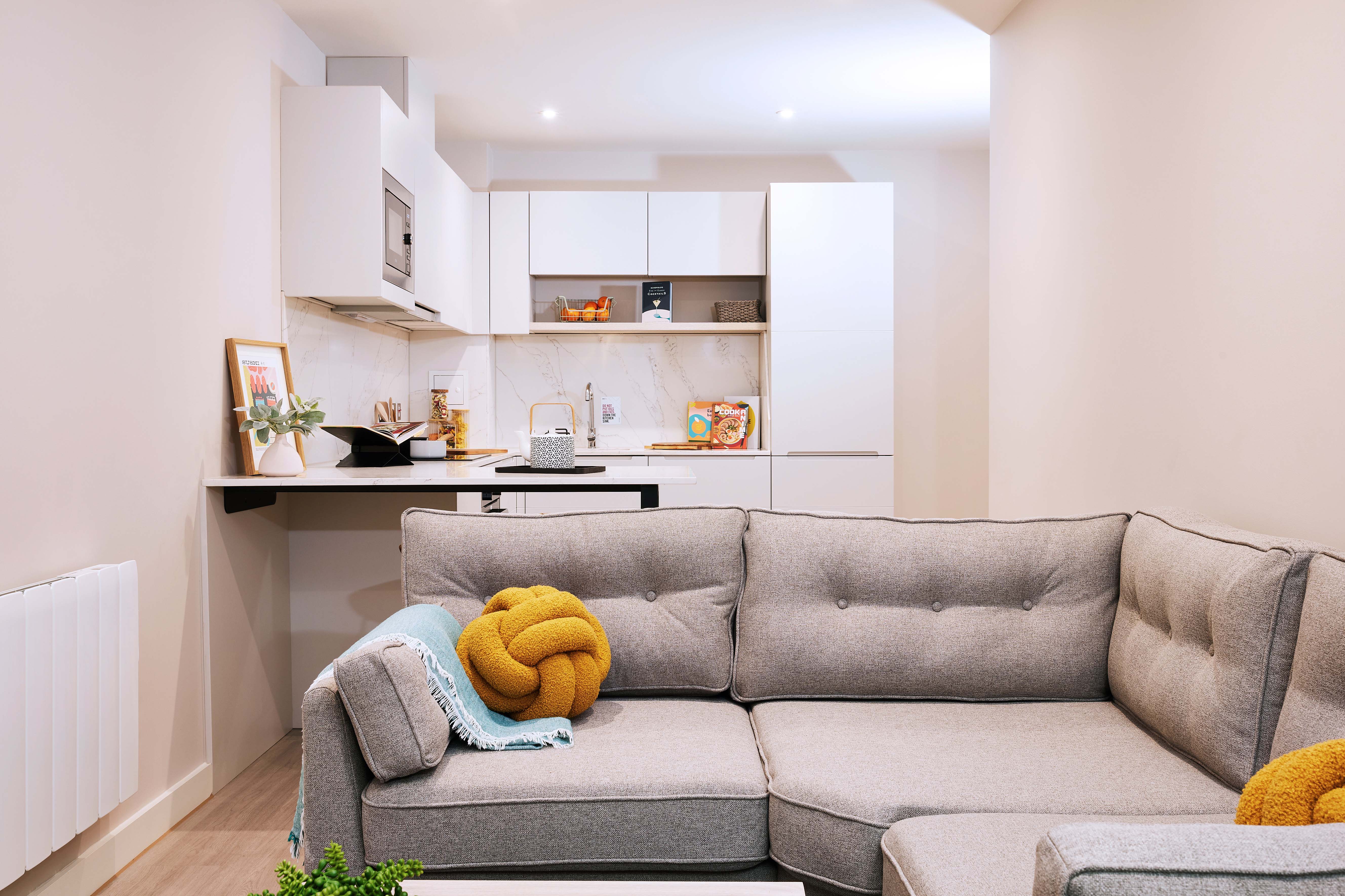 A modern living room features a gray L-shaped sofa with colorful cushions, a small coffee table, and a compact kitchen in the background. The kitchen has white cabinets, a counter with plants, and some decorative items.