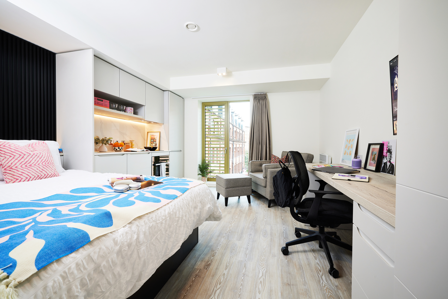 A modern bedroom on Bruce Street features a bed with a blue and white blanket, a desk with a chair, laptop, and books to the right. A cozy sitting area near the glass door leads to a balcony. Light wood flooring runs throughout the room.