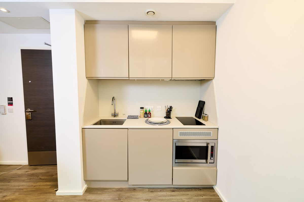 Compact modern kitchenette with beige cabinets, sink, stove, microwave, utensils, and condiments on the countertop; set against a white wall with wooden flooring.