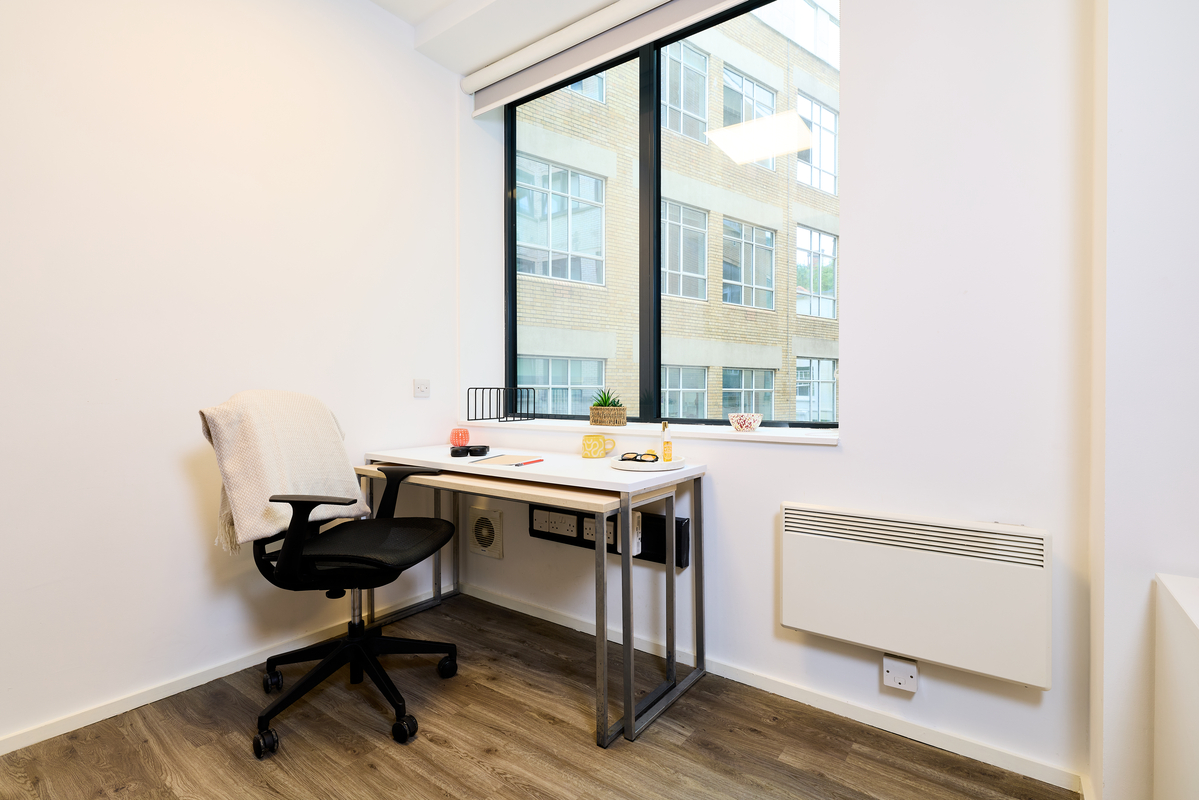 A minimalist office space with a black swivel chair at a white desk by a large window. The desk has a laptop, mug, plant, notepad, and glasses. Wooden floor and white walls create a clean, bright atmosphere.