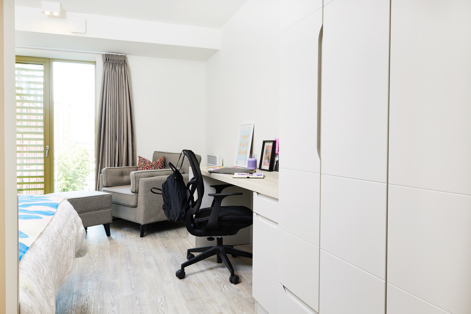 A bright, modern bedroom on Bruce Street features a bed on the left, a gray armchair with a footrest, and a desk with an office chair. The desk holds books and a framed picture. A large wardrobe sits on the right, and there's a window with curtains in the background.