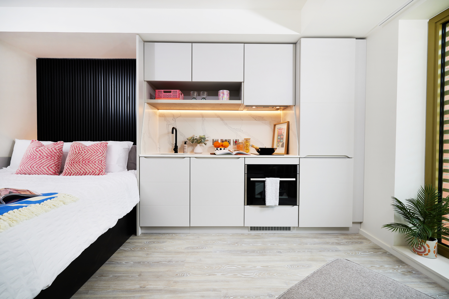 A modern studio apartment on Bruce Street featuring a compact kitchen with white cabinets, black fixtures, and a marble backsplash. A cozy bed with pink pillows is adjacent. The space is bright, with wooden flooring and a small plant by the window.