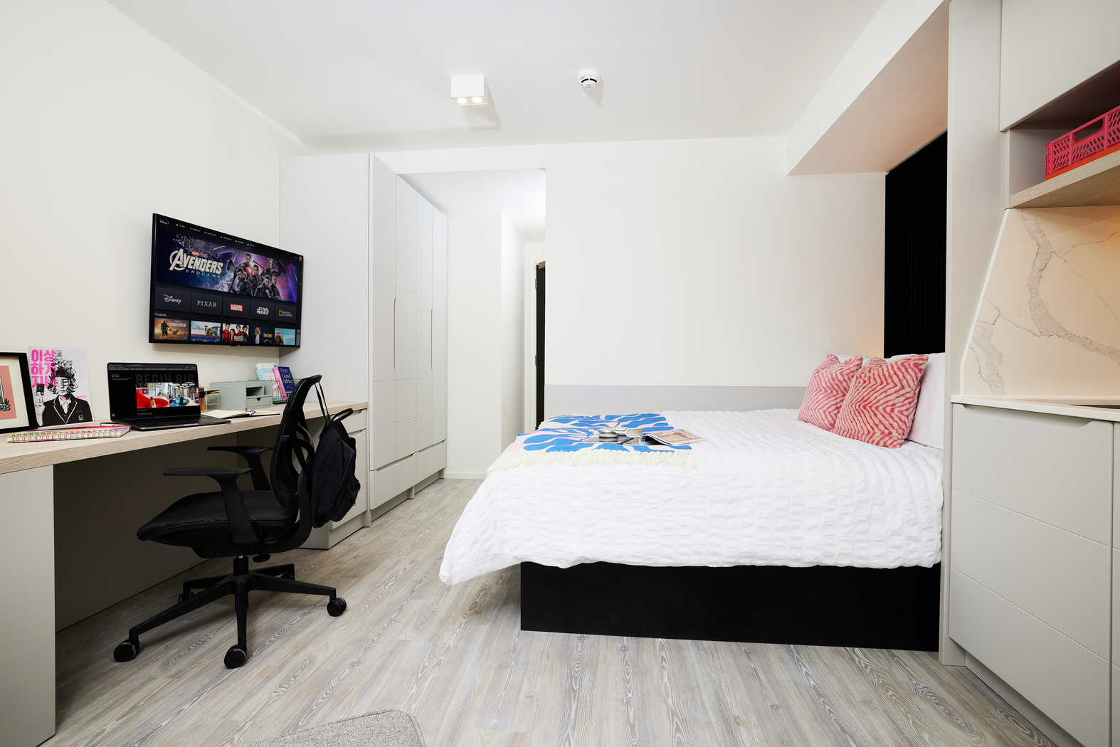 A modern bedroom on Bruce Street features a bed with decorative pillows and a magazine atop the comforter. A desk holds a computer streaming your favorite shows, accompanied by a keyboard and black chair. The room boasts wooden flooring, white walls, and sleek built-in cabinets.