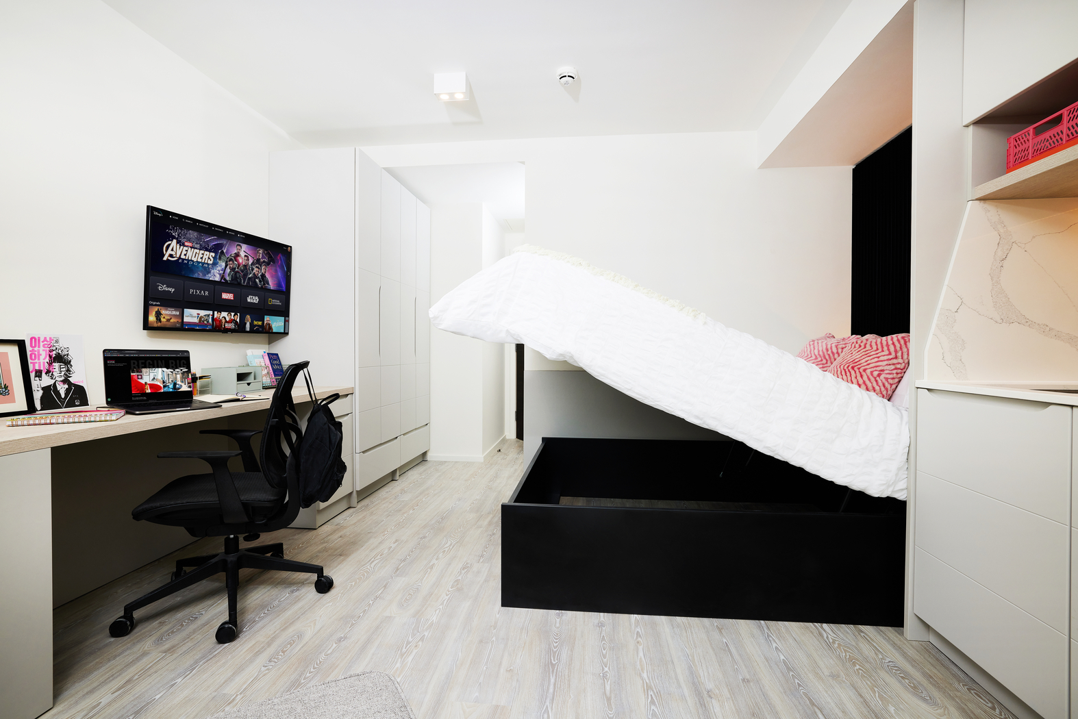 A compact, modern bedroom on Bruce Street features a bed that lifts for storage, revealing an empty compartment. A desk with a computer and chair is on the left, while a large TV mounted on the wall displays a streaming service. The room boasts light wood flooring, adding warmth to the sleek design.
