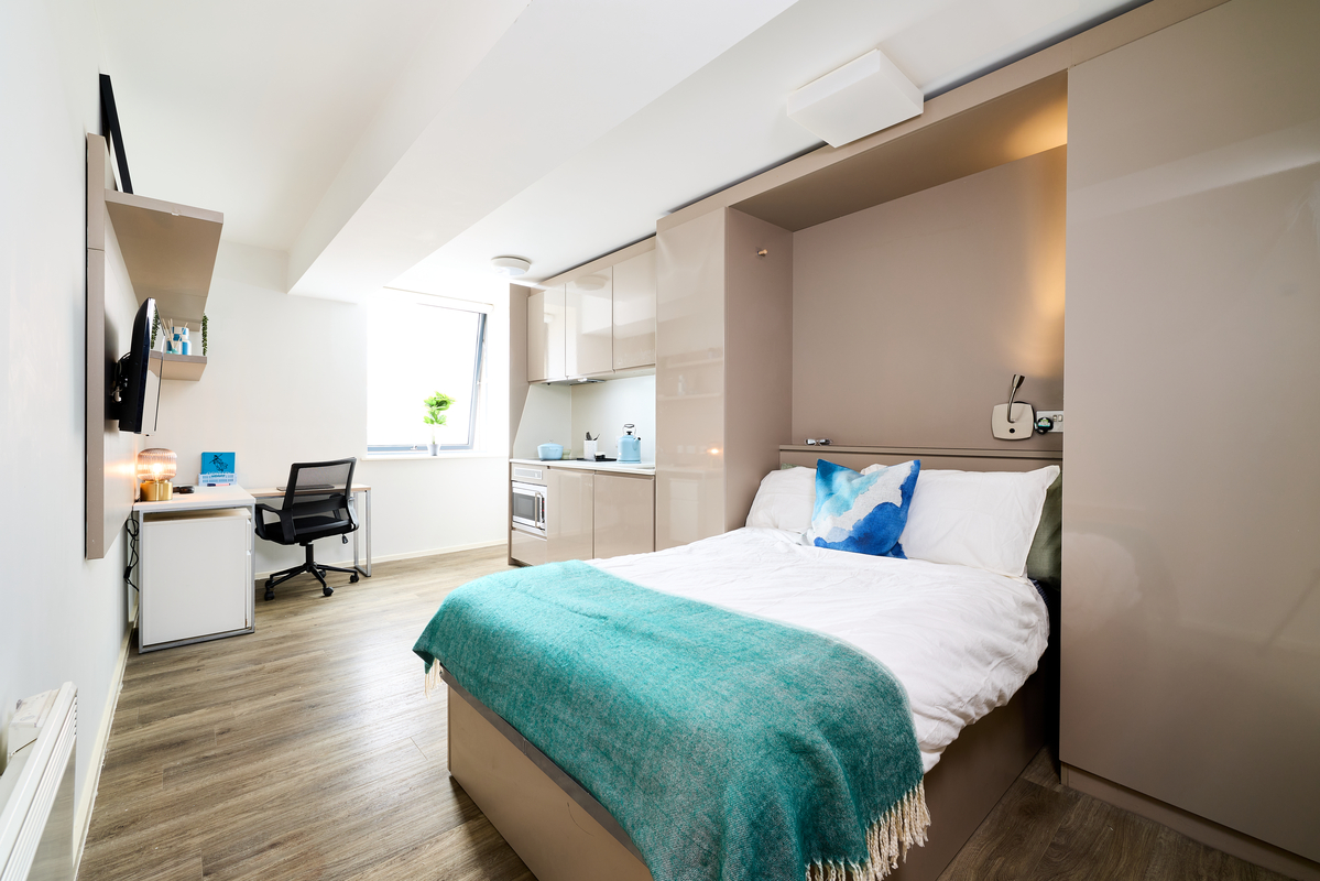 Modern studio apartment in Portland House with a bed, turquoise blanket, white bedding, small kitchen area, desk with a chair by a window, and light wood flooring. The room is bright and neatly organized.