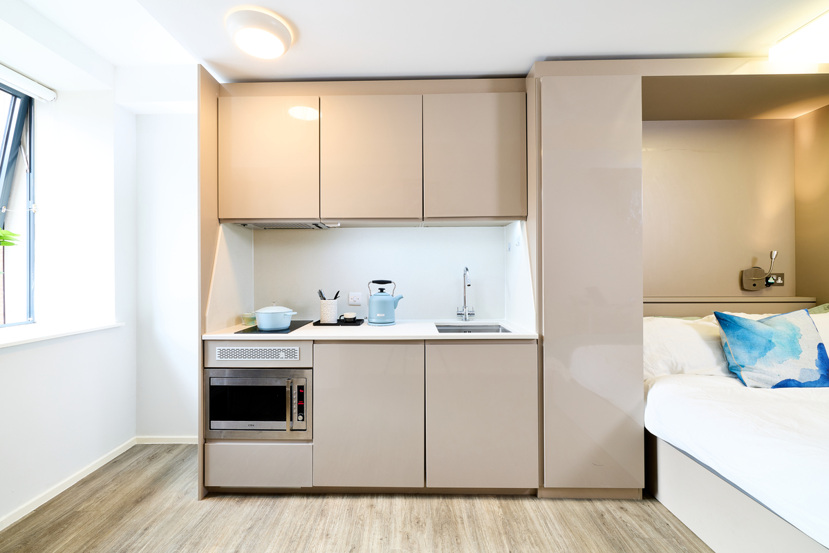 Modern studio apartment kitchen at Portland House with beige cabinets, built-in microwave, stove, blue kettle, and cookware beside a cozy bed with white bedding and blue pillows, all brightly lit near a window.