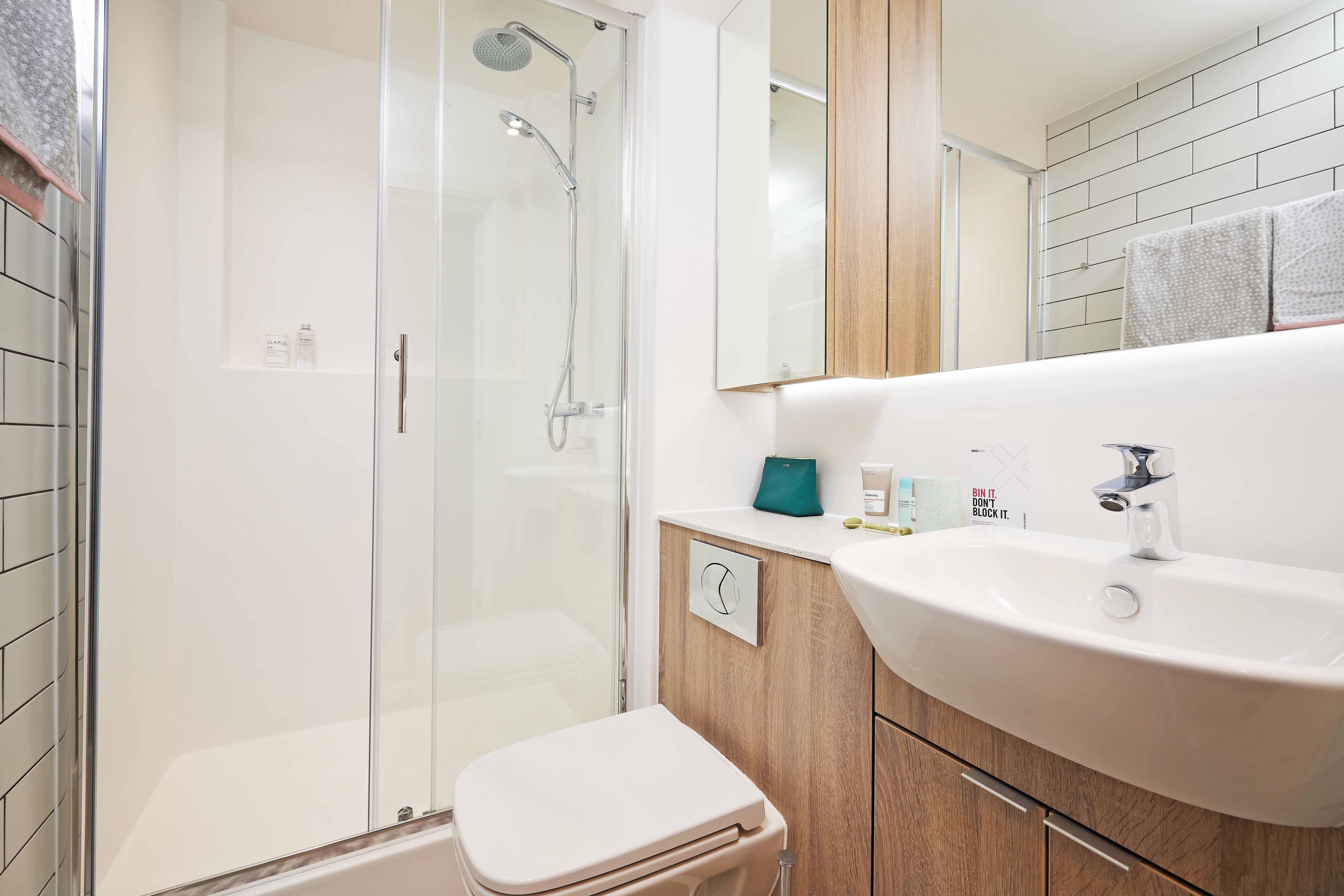 Modern bathroom with glass-enclosed shower, white sink, wooden cabinets, and wall-mounted toilet. The walls are tiled with white tiles, and towels are neatly arranged on the shelf. A small teal item sits on the countertop.