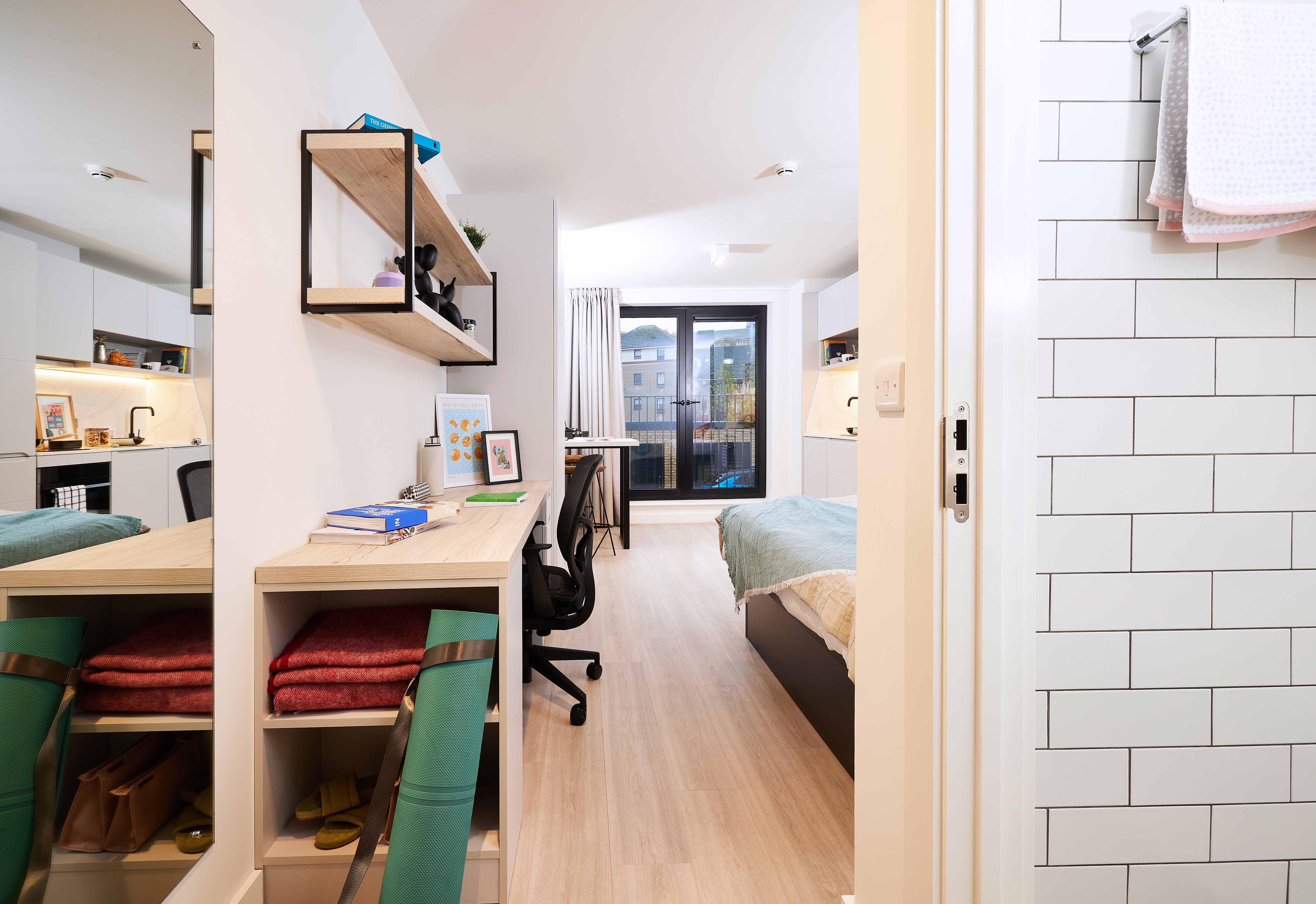 Modern studio apartment with light wooden flooring. Features a compact kitchen, desk with a chair, shelving, and a bed near a large window. Towels hang by a bathroom entrance with white subway tiles on the wall. Cozy and bright ambiance.