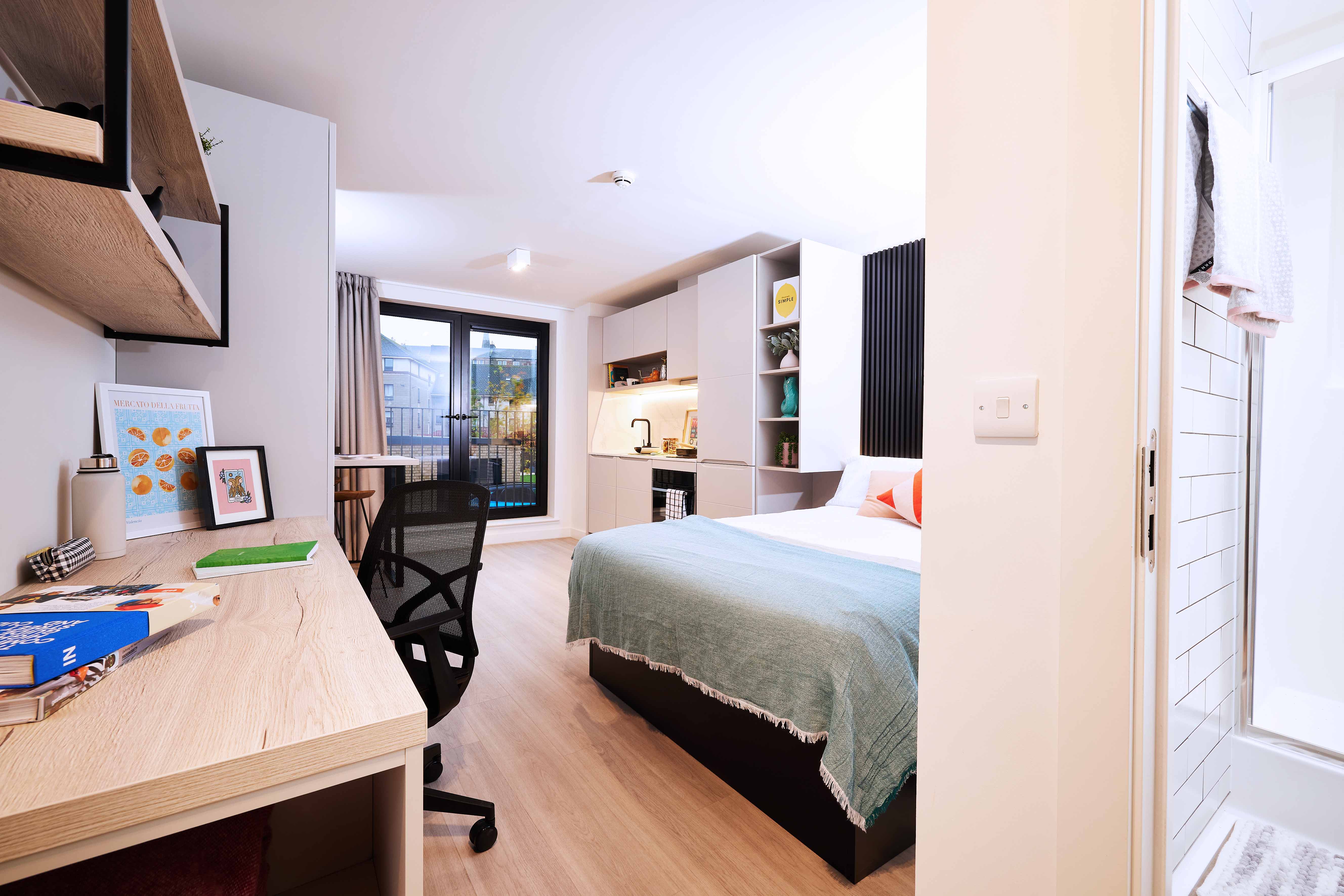 Modern bedroom with wooden flooring, featuring a bed with a green throw, a desk with books and a laptop, shelves, a black chair, and a kitchenette. A glass door opens to a balcony, and a door leads to a bathroom.