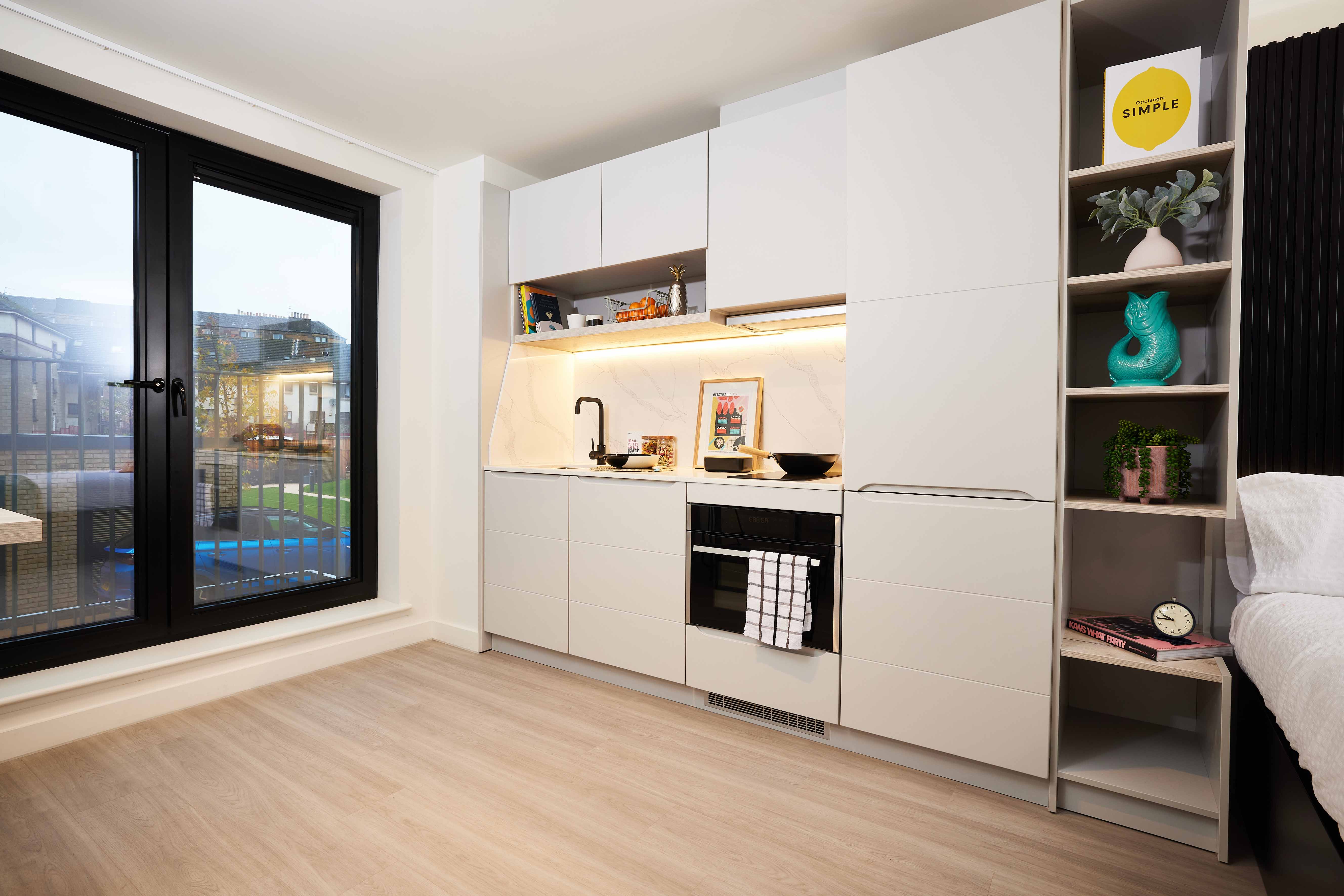 Modern studio apartment with light wood flooring. Features a sleek kitchenette with white cabinets, microwave, and sink. Open shelving displays decor including a plant and teal statue. Large windows on the left offer a view of a courtyard.