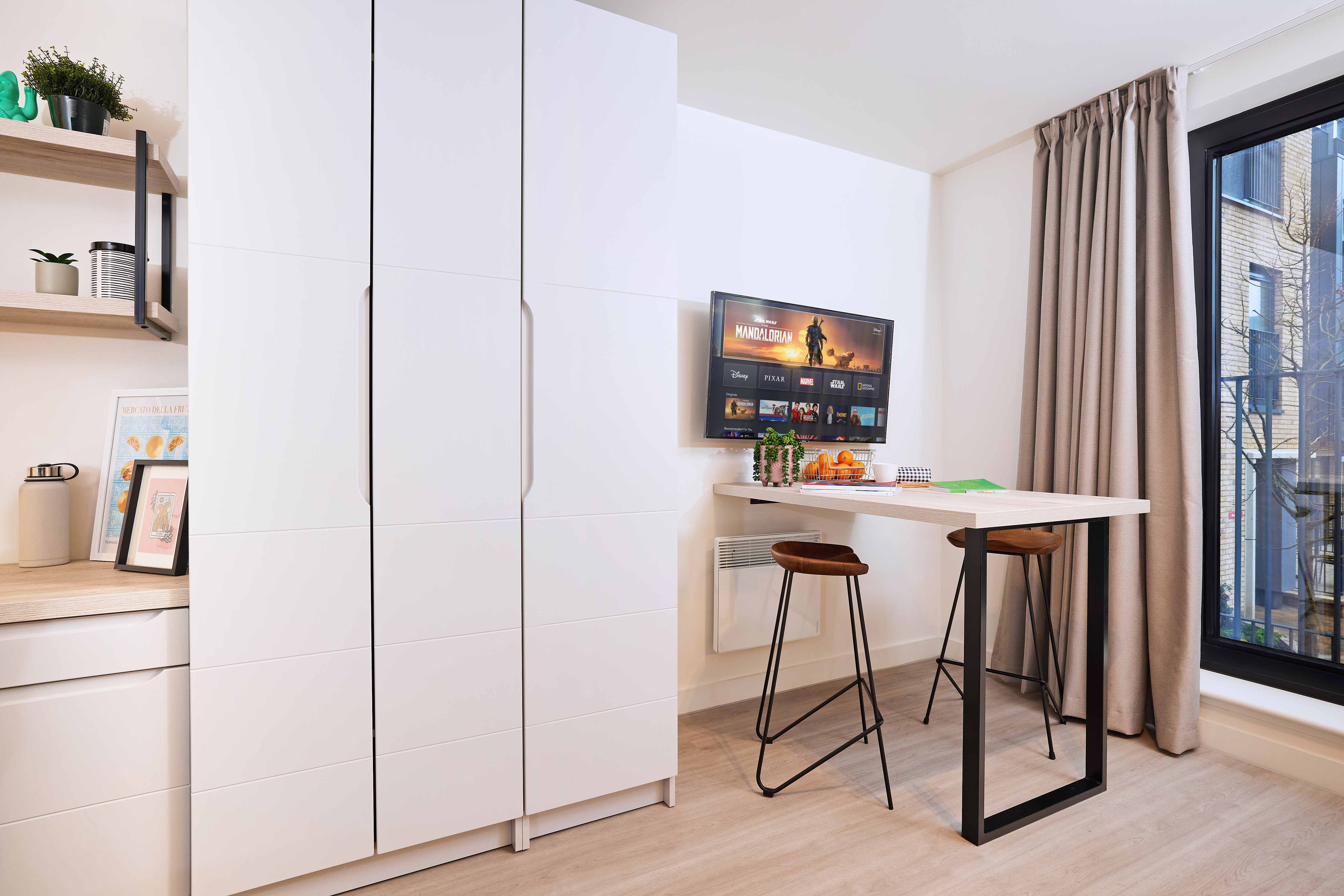 Modern kitchen with white cabinets, a wall-mounted TV displaying a game, and a high table with two wooden barstools. Shelves hold plants and books. A large window with beige curtains allows natural light to fill the space.