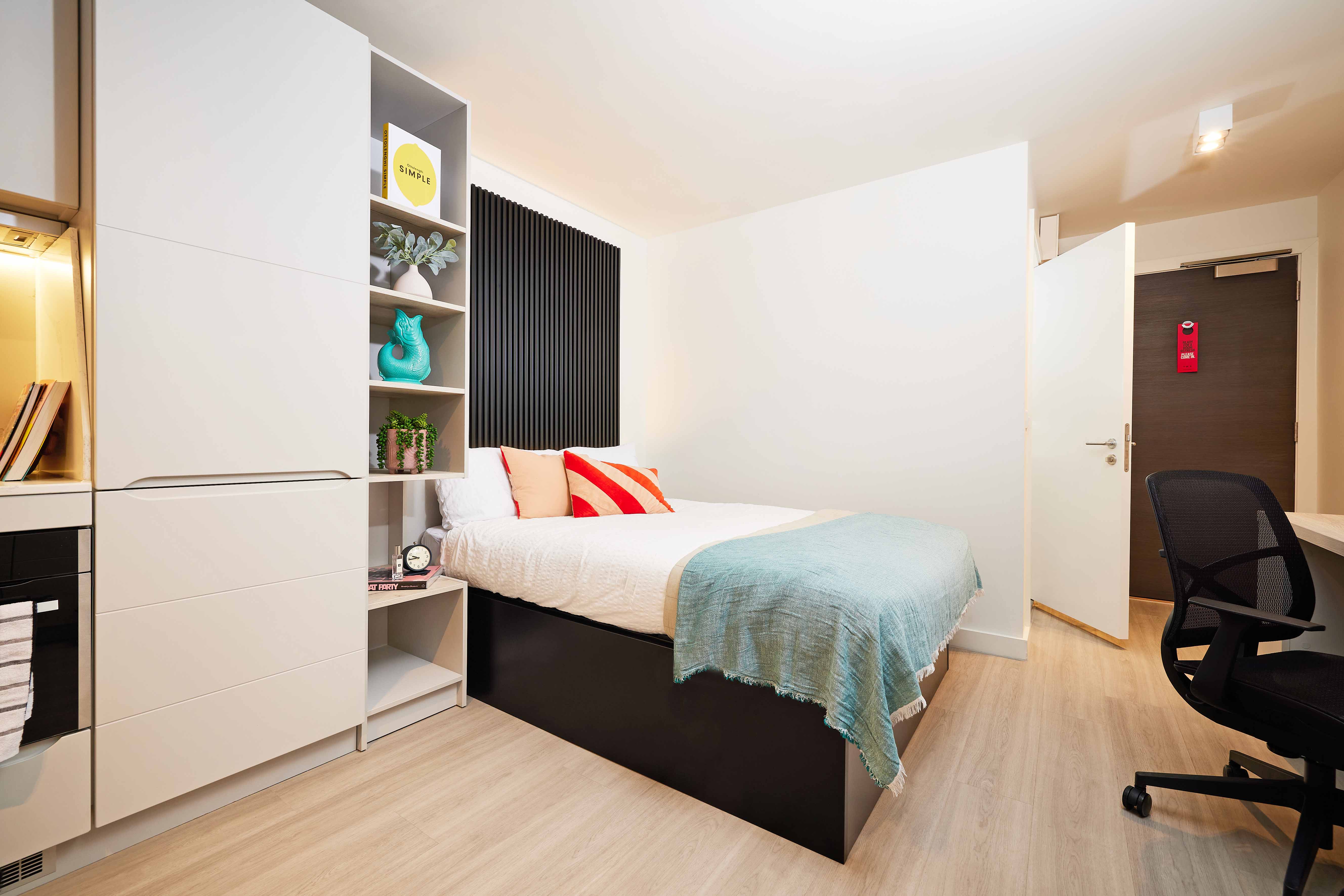 Modern bedroom with light wood flooring, a bed with white and teal bedding, and colorful pillows. A shelving unit holds books and decorations. There's a chair and desk on the right and a dark door in the background with a red sign.