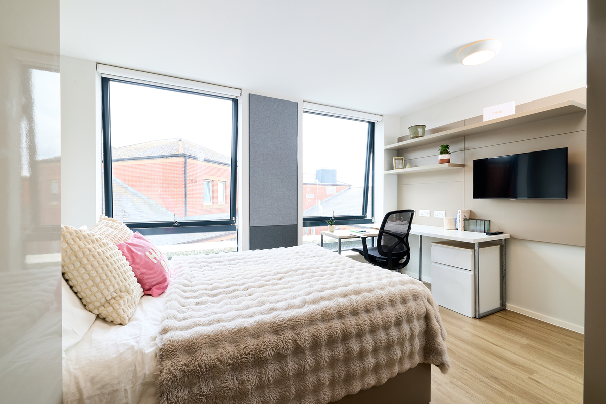 A bright, modern Portland House bedroom features a large bed with textured bedding, a desk with a chair by tall windows, shelves with decor, a wall-mounted TV, and light wood flooring.