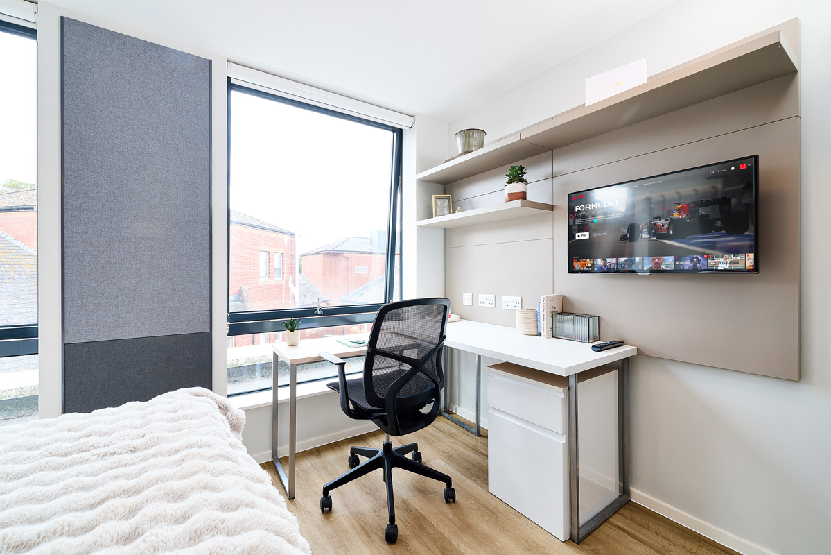 A modern, tidy bedroom at Portland House features a single bed, a large window, a white desk and chair, shelves with decor, and a wall-mounted TV displaying a streaming service interface.