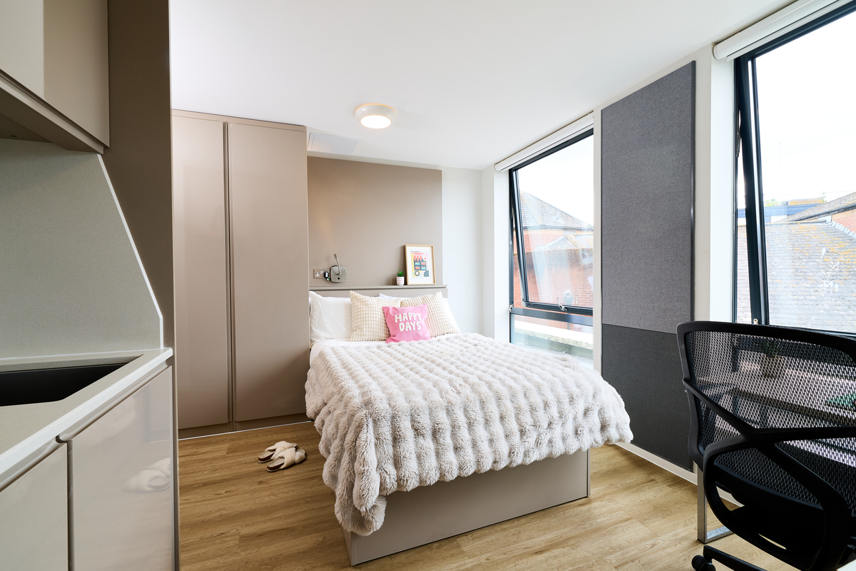 A bright, modern bedroom at Portland House with a large window, a neatly made bed with a fluffy white blanket, a "HAPPY DAYS" cushion, wooden floor, wardrobe, and a black office chair by the desk.