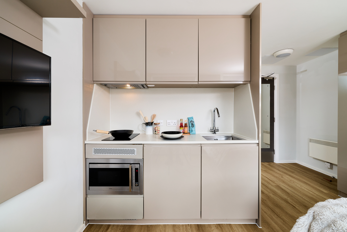 A compact modern kitchen in Portland House with beige cabinets, a sink, an electric cooktop with a wok, utensils, some food items, a microwave, and wood flooring. A TV is mounted on the wall to the left.