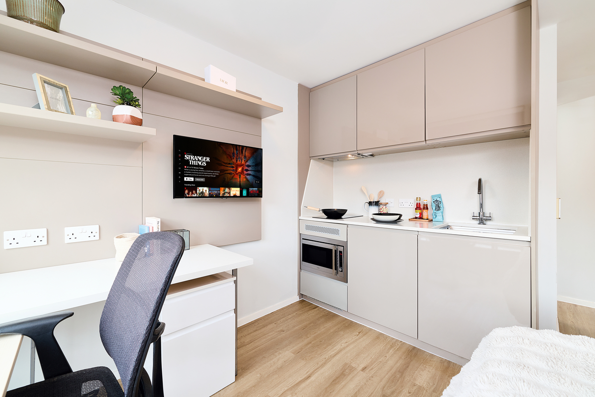 Modern studio apartment in Portland House with a compact kitchen area, desk and chair, wall-mounted TV displaying "Stranger Things," light wood flooring, plus shelves holding decor items and plants.